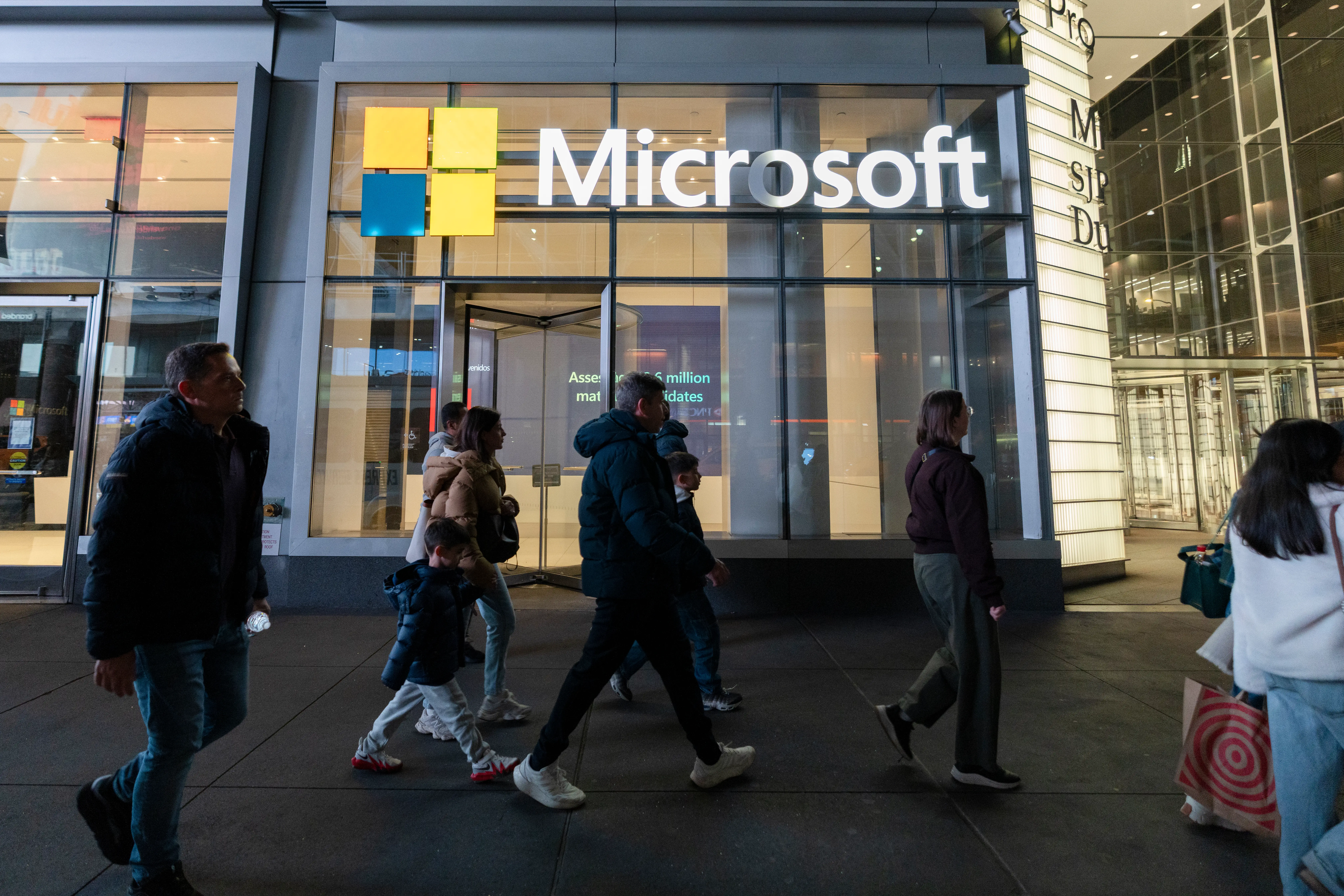 Pedestrians walk past a Microsoft storefront with illuminated signage and logo at dusk.