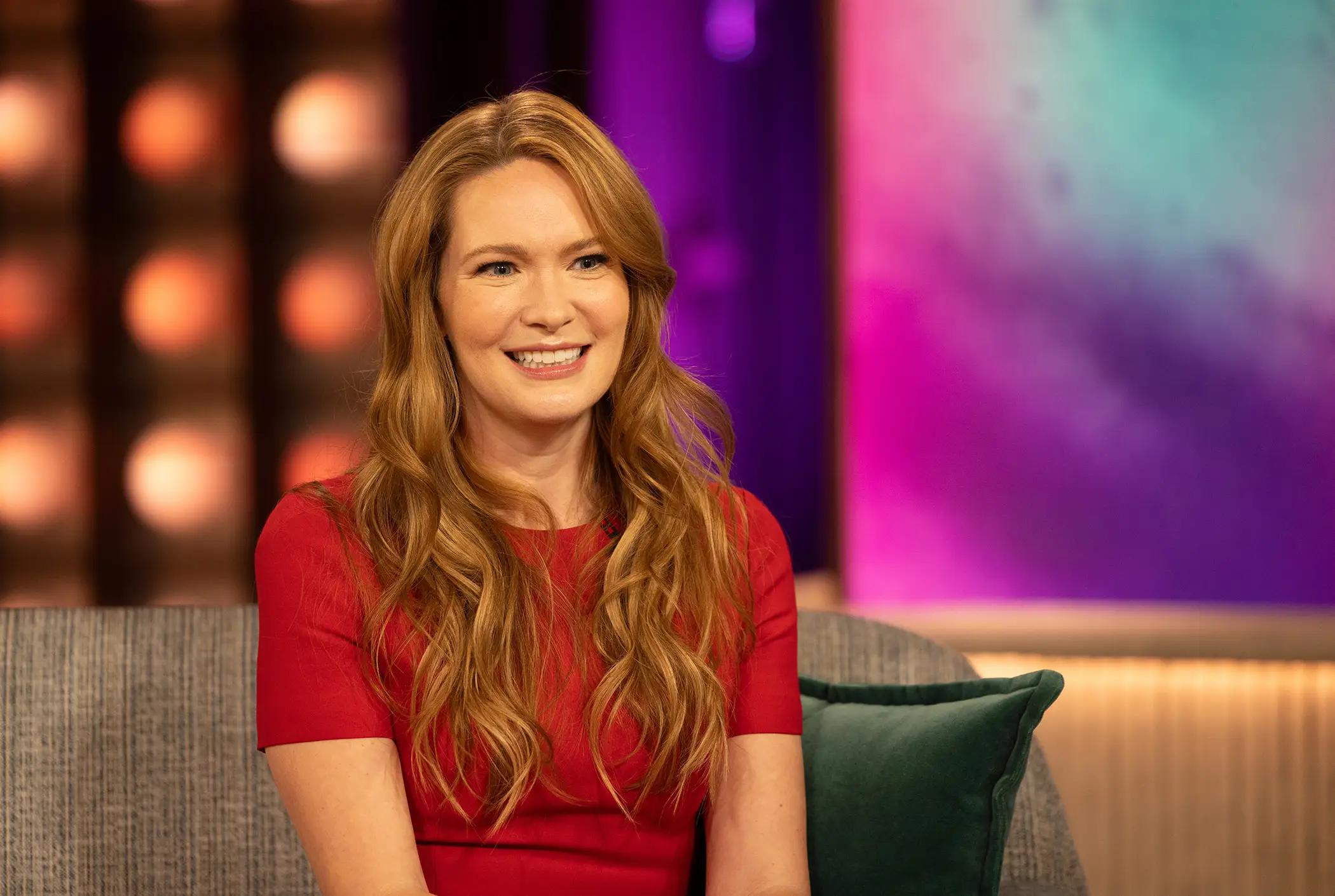 Sarah J. Maas in a red dress sits on a studio couch against colorful purple and teal lighting.