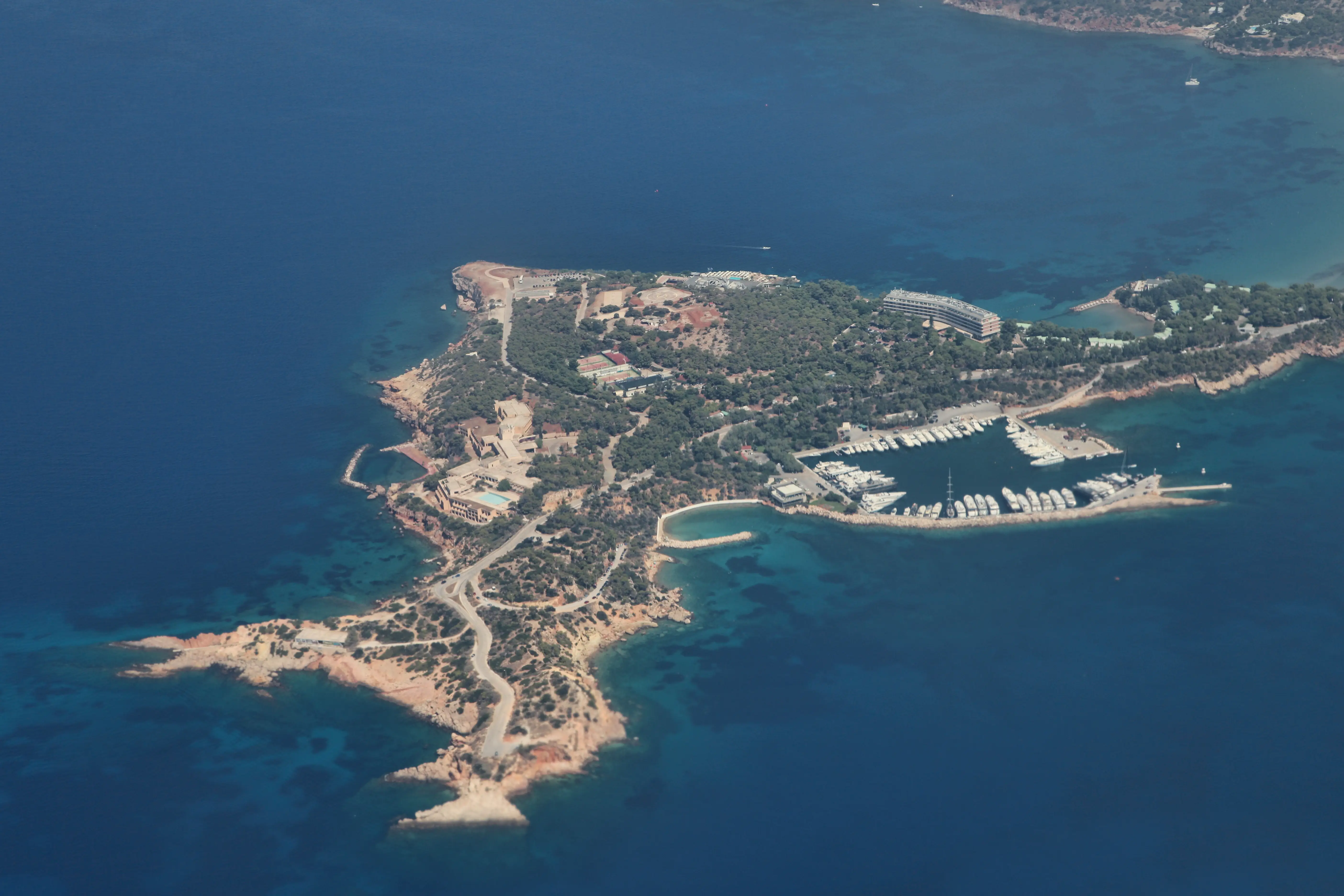 Aerial view of a rocky coastal peninsula with marina, buildings, roads, and deep blue surrounding sea.