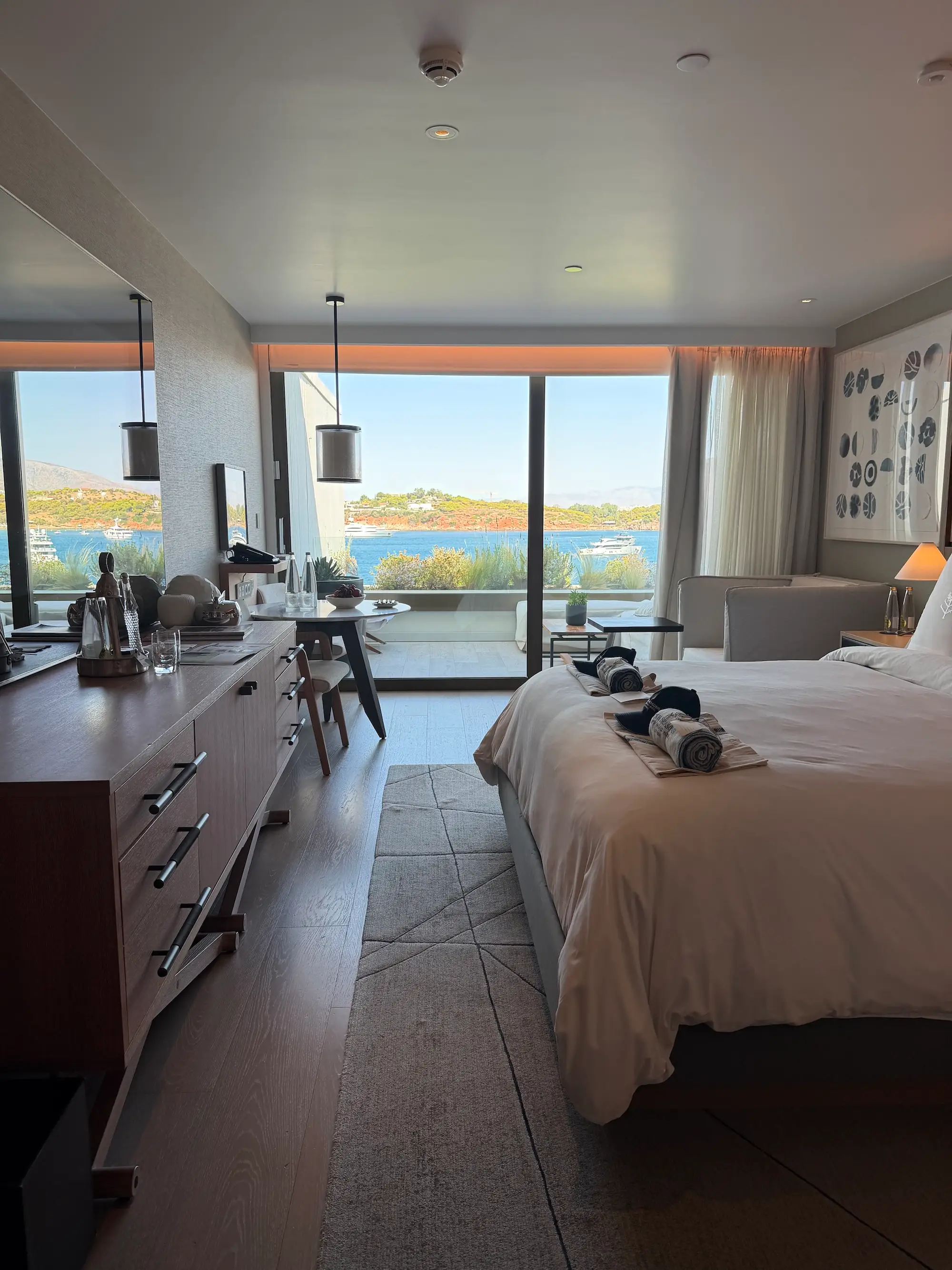 Modern hotel room with a large bed, desk, and balcony overlooking a blue waterfront and hills.