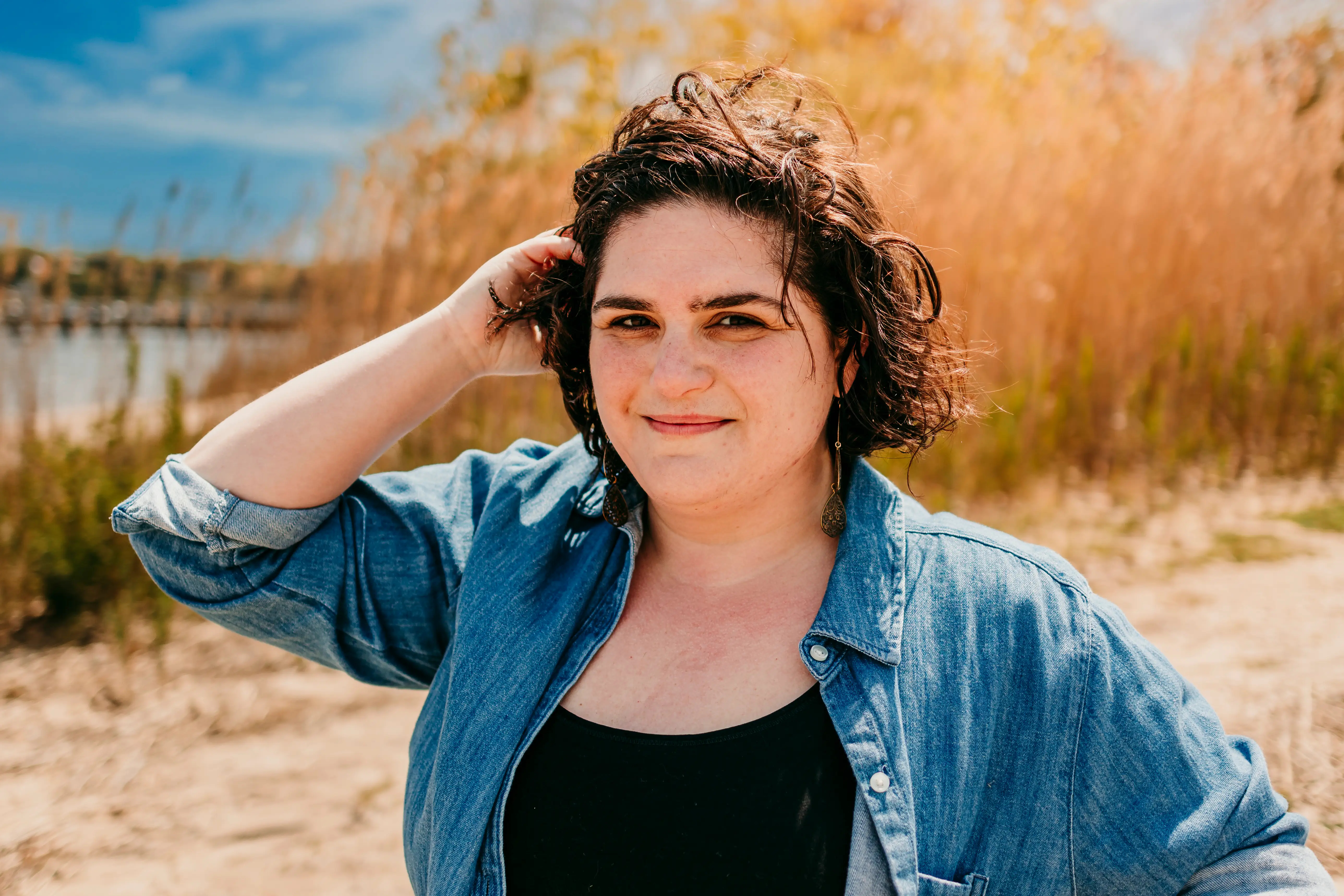 Person in denim shirt smiles outdoors near tall reeds and water on a sunny day.