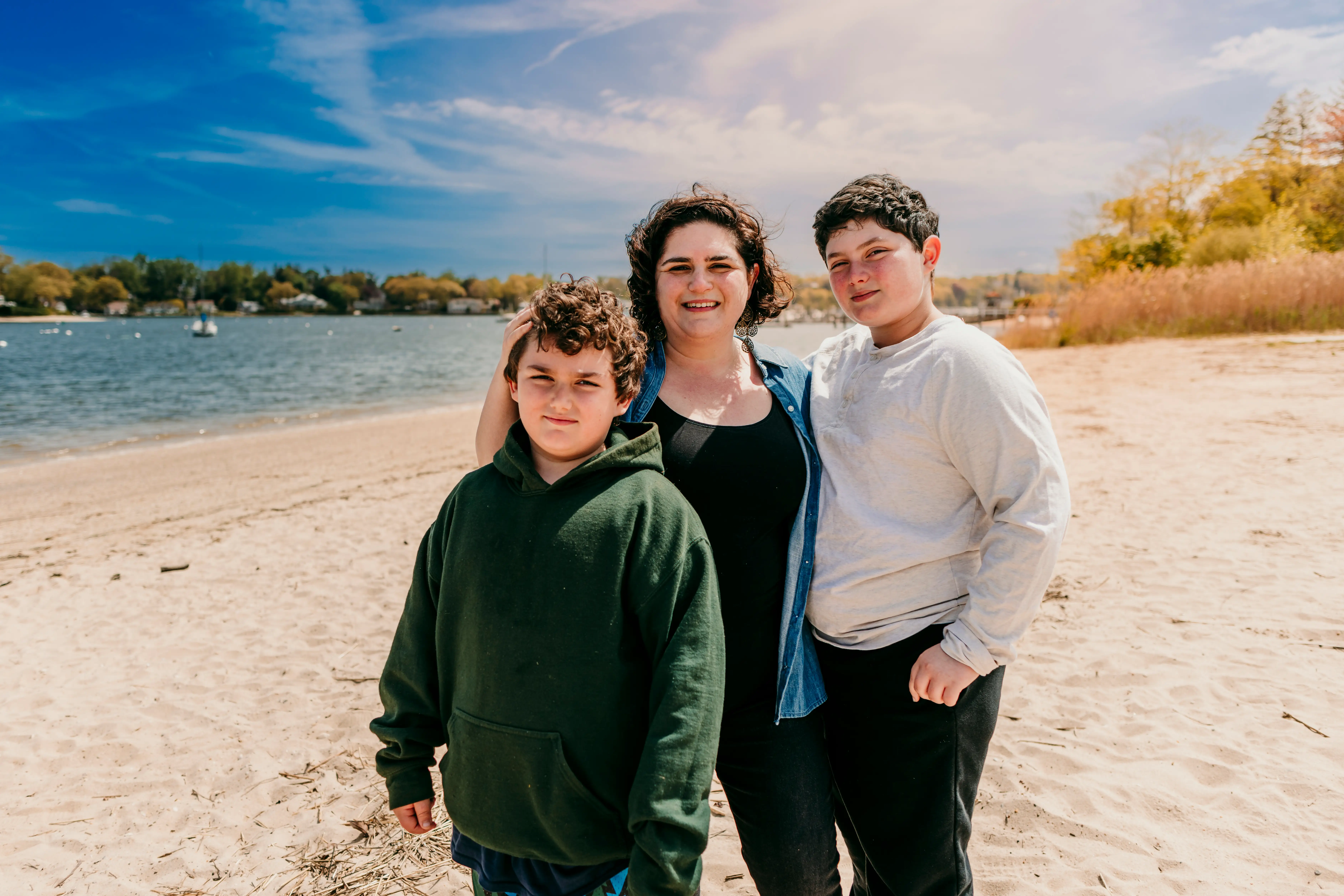 Three people stand together on a sandy beach beside a lake under a partly cloudy sky.
