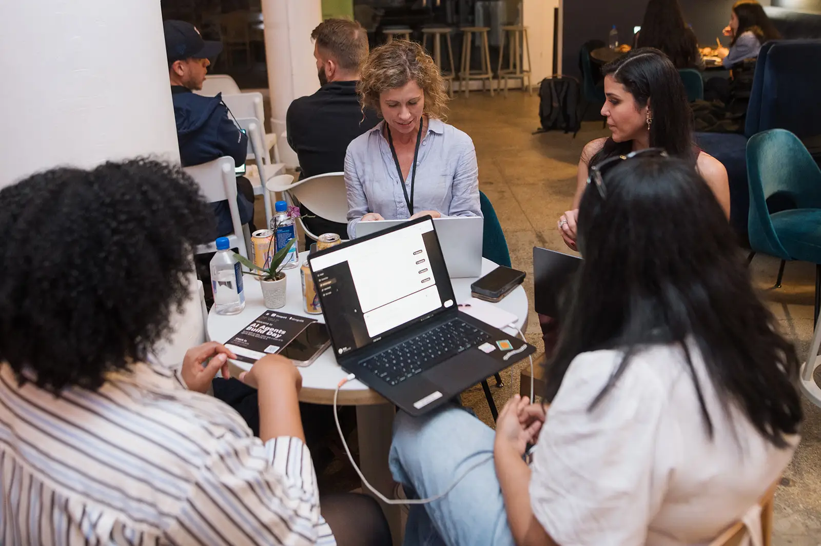 Five people sit around a small table with laptops and drinks in a cafe workspace.