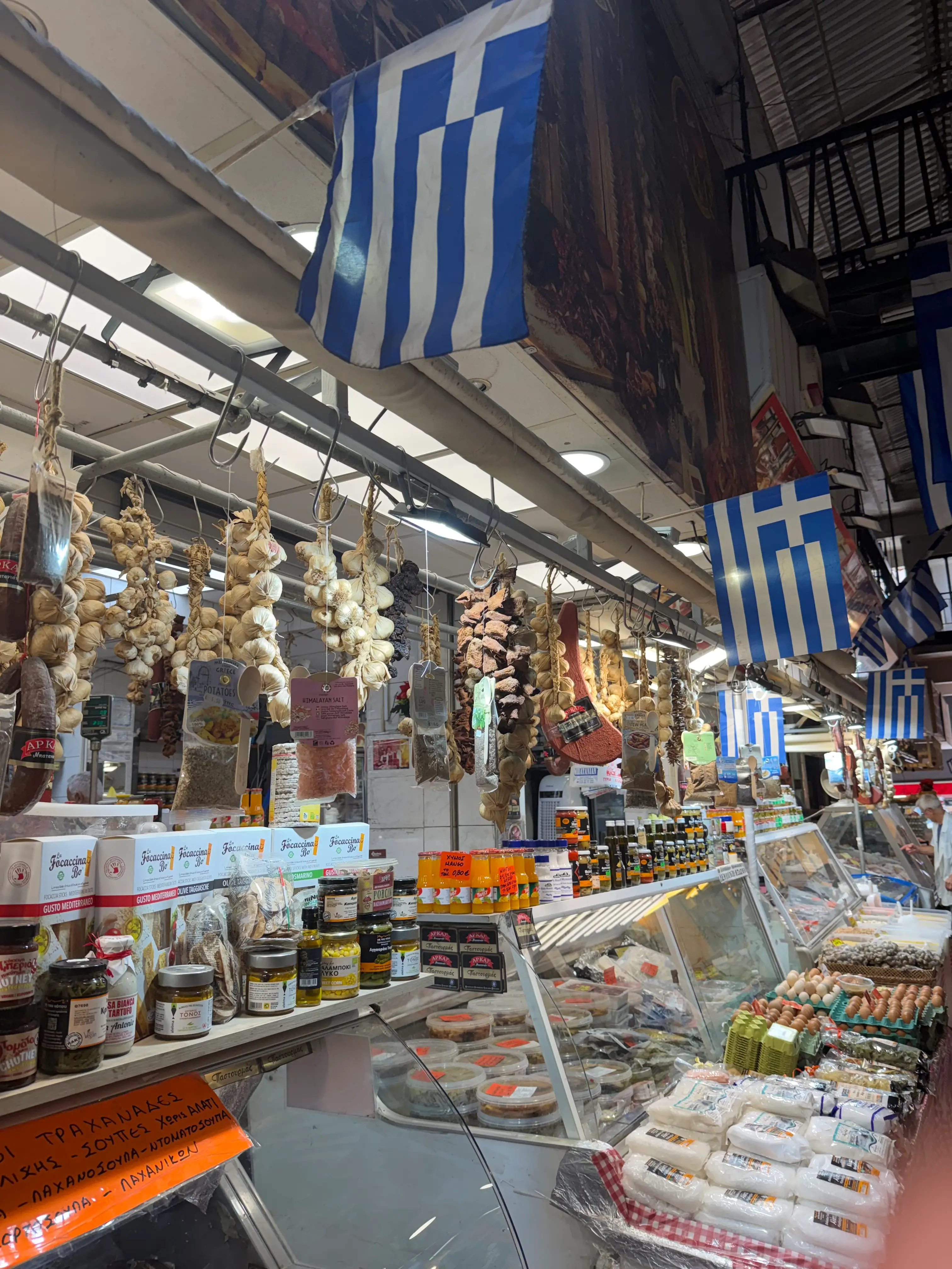 Greek flags hang above a market deli counter with garlic strings, packaged foods, eggs, and refrigerated goods.