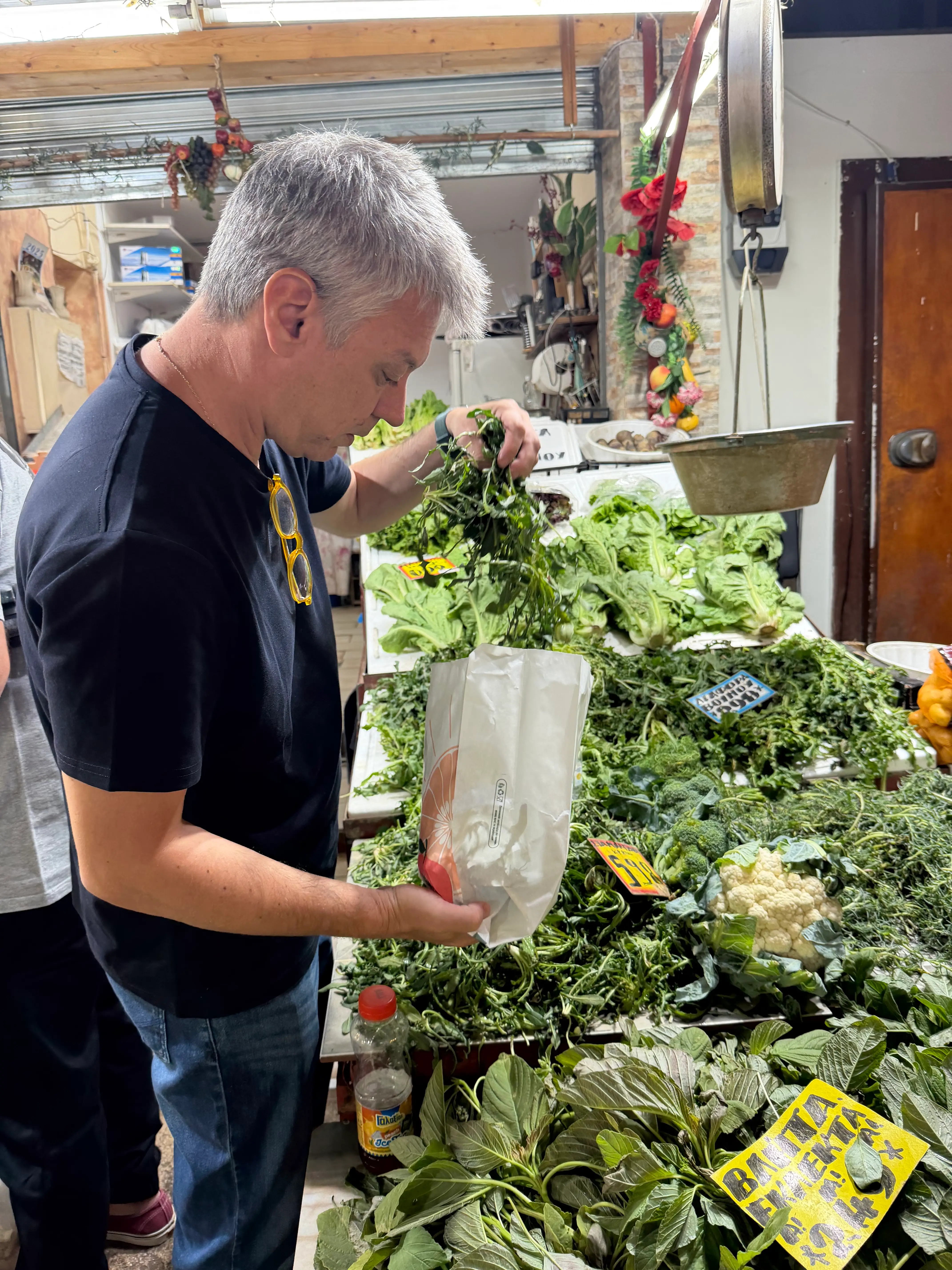 Shopper places fresh leafy greens into a paper bag at a crowded produce market stall.