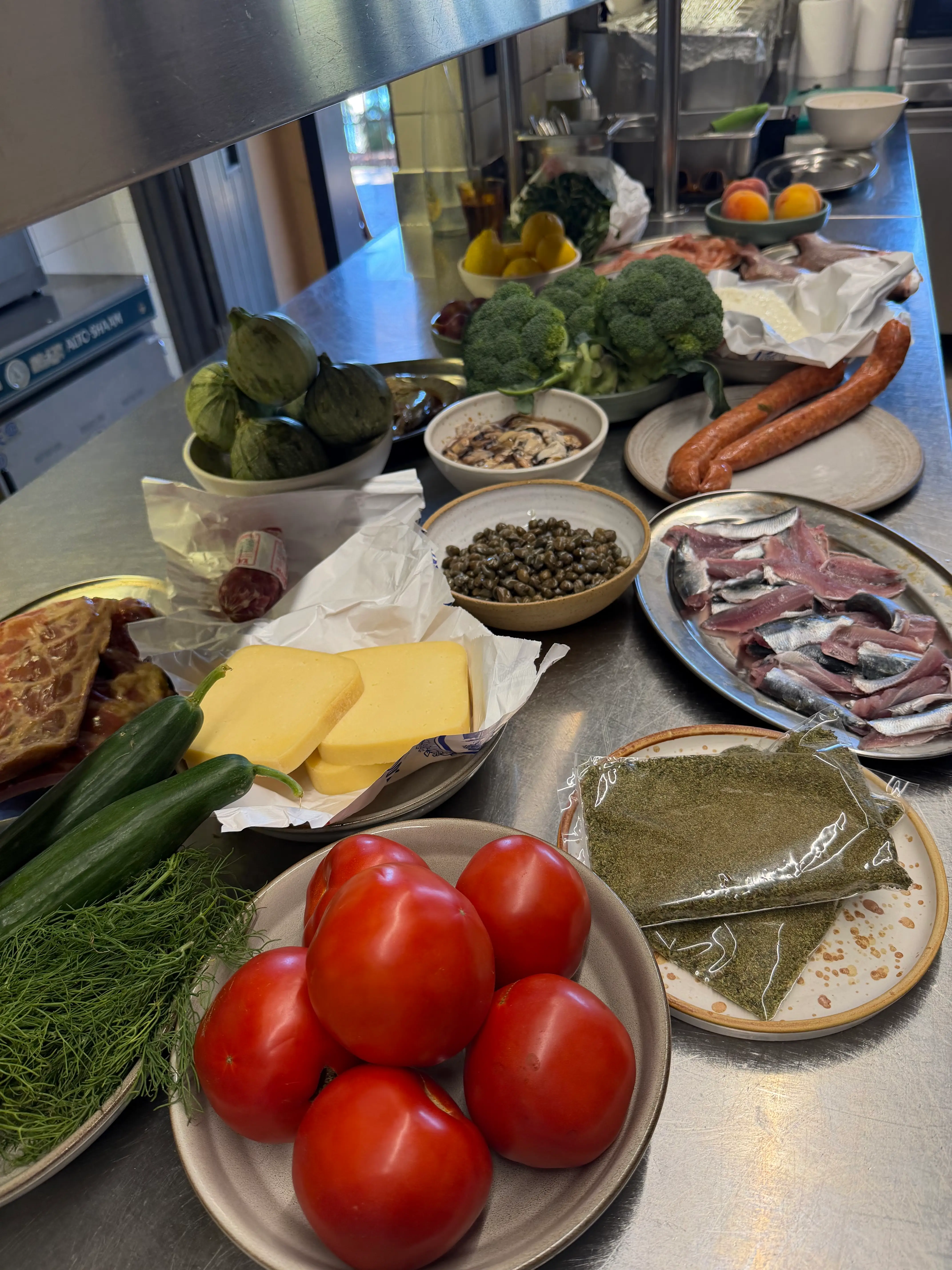 Assorted vegetables, fish, cheese, sausages, herbs, and capers are arranged on a stainless-steel kitchen counter.