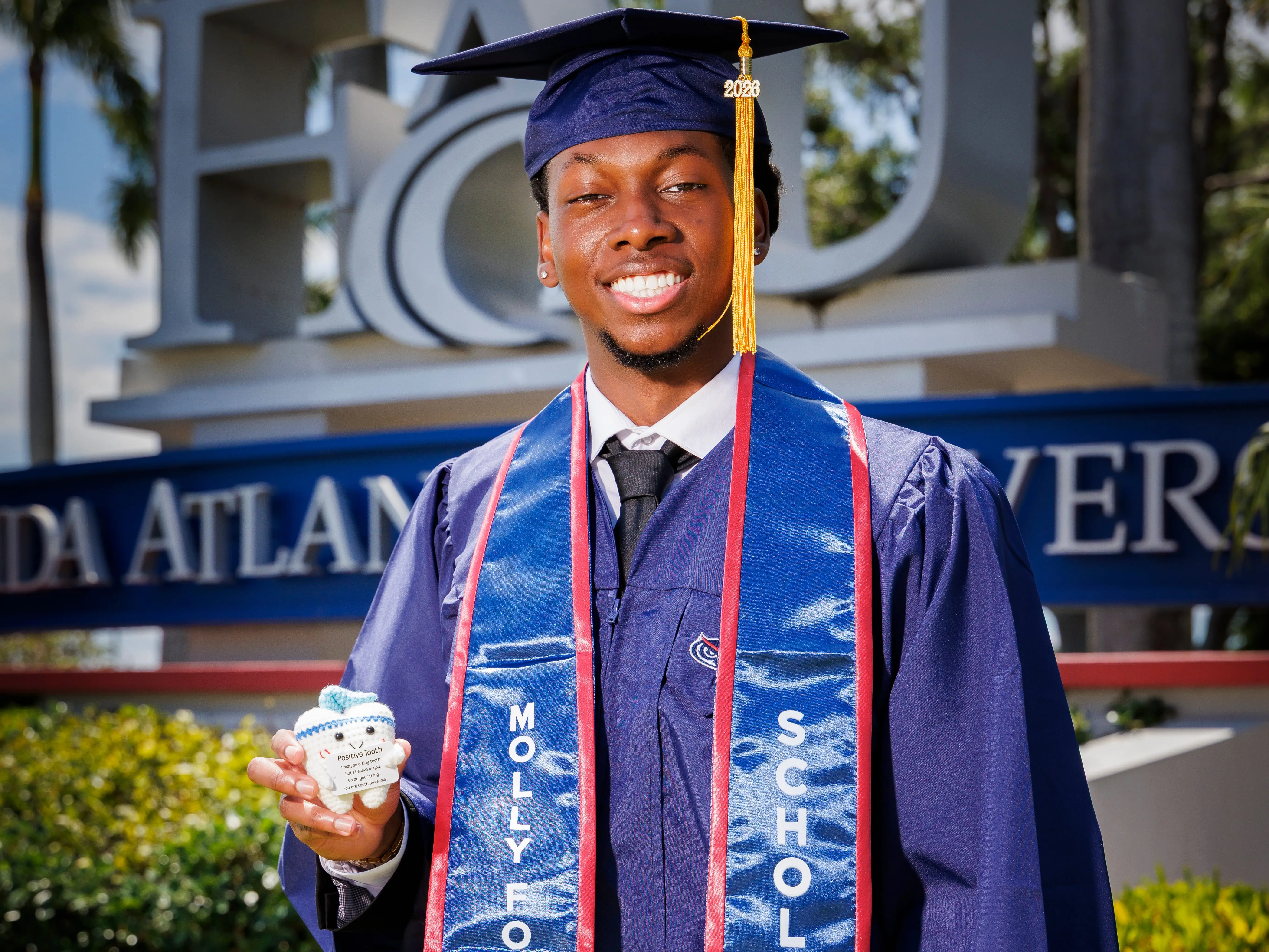 Graduate in navy cap and gown poses by a FAU sign while wearing a blue Molly Ford Scholar stole.