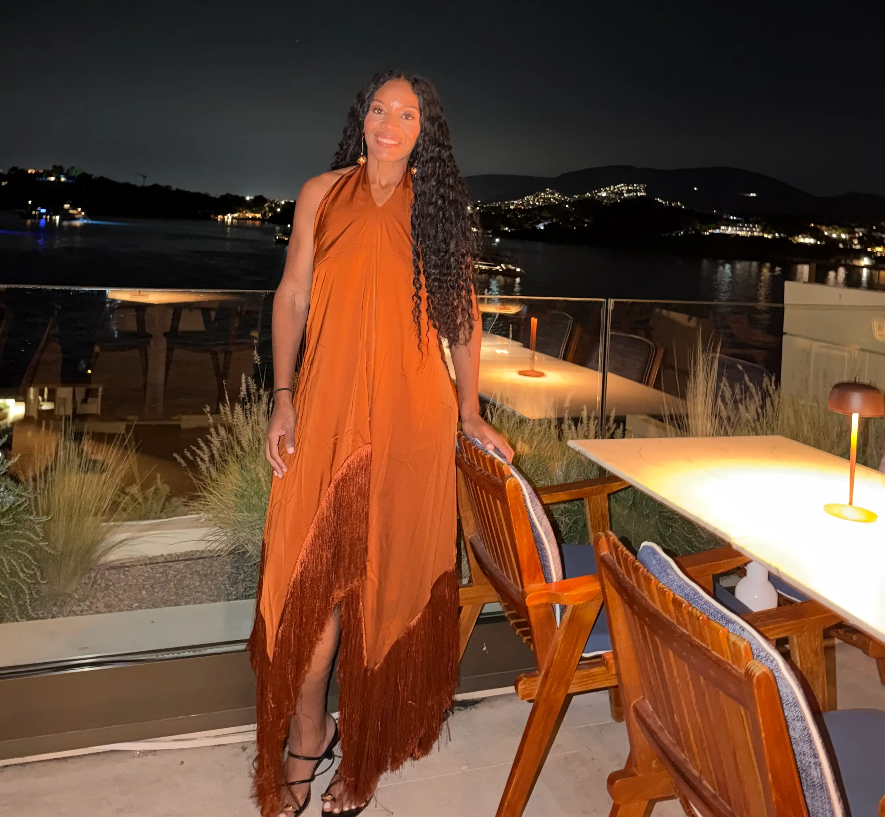 Person in a rust-orange fringed dress stands on a nighttime waterfront terrace with illuminated tables and bay views.