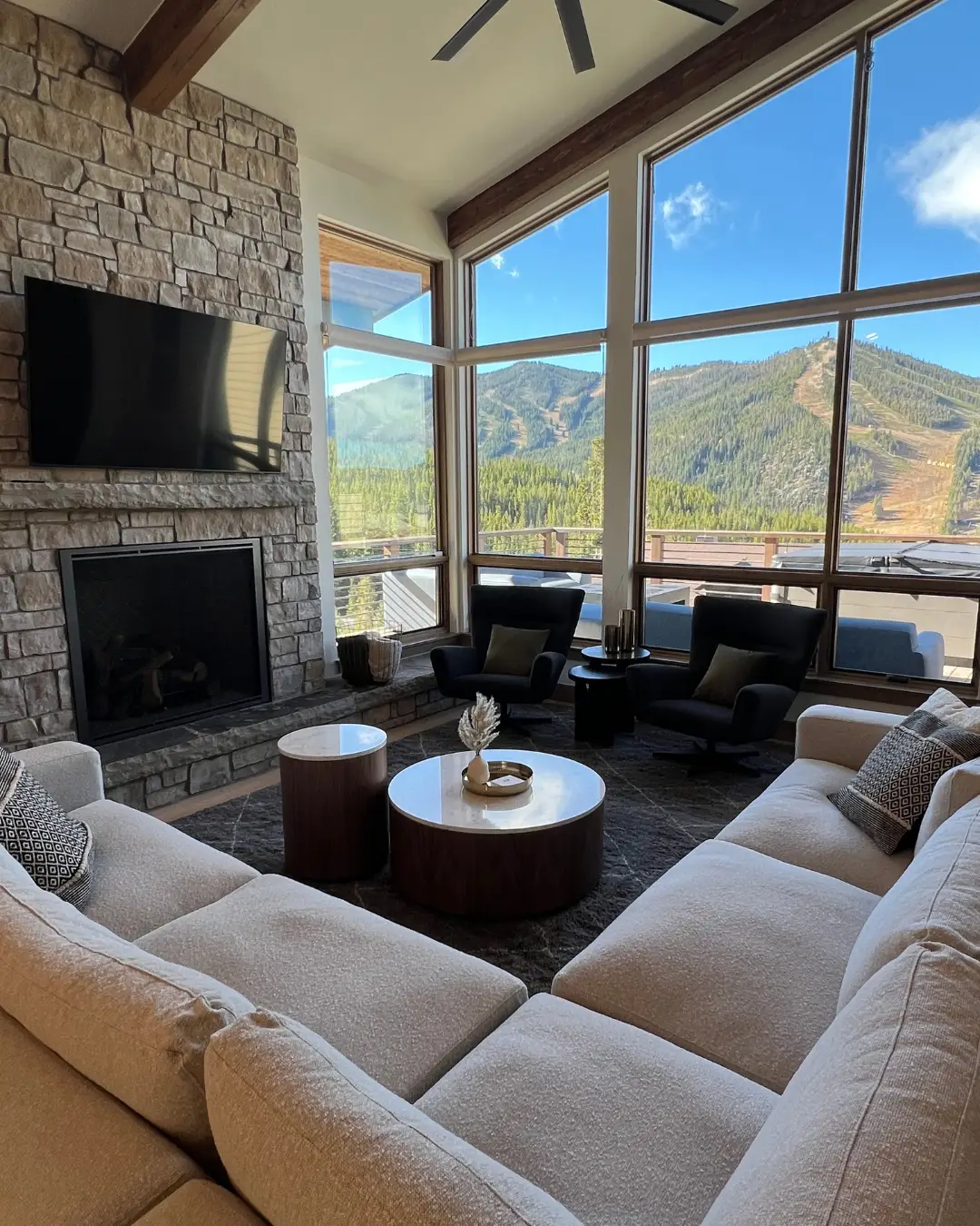 Modern living room with stone fireplace, large windows, sectional seating, and mountain views beyond a balcony.
