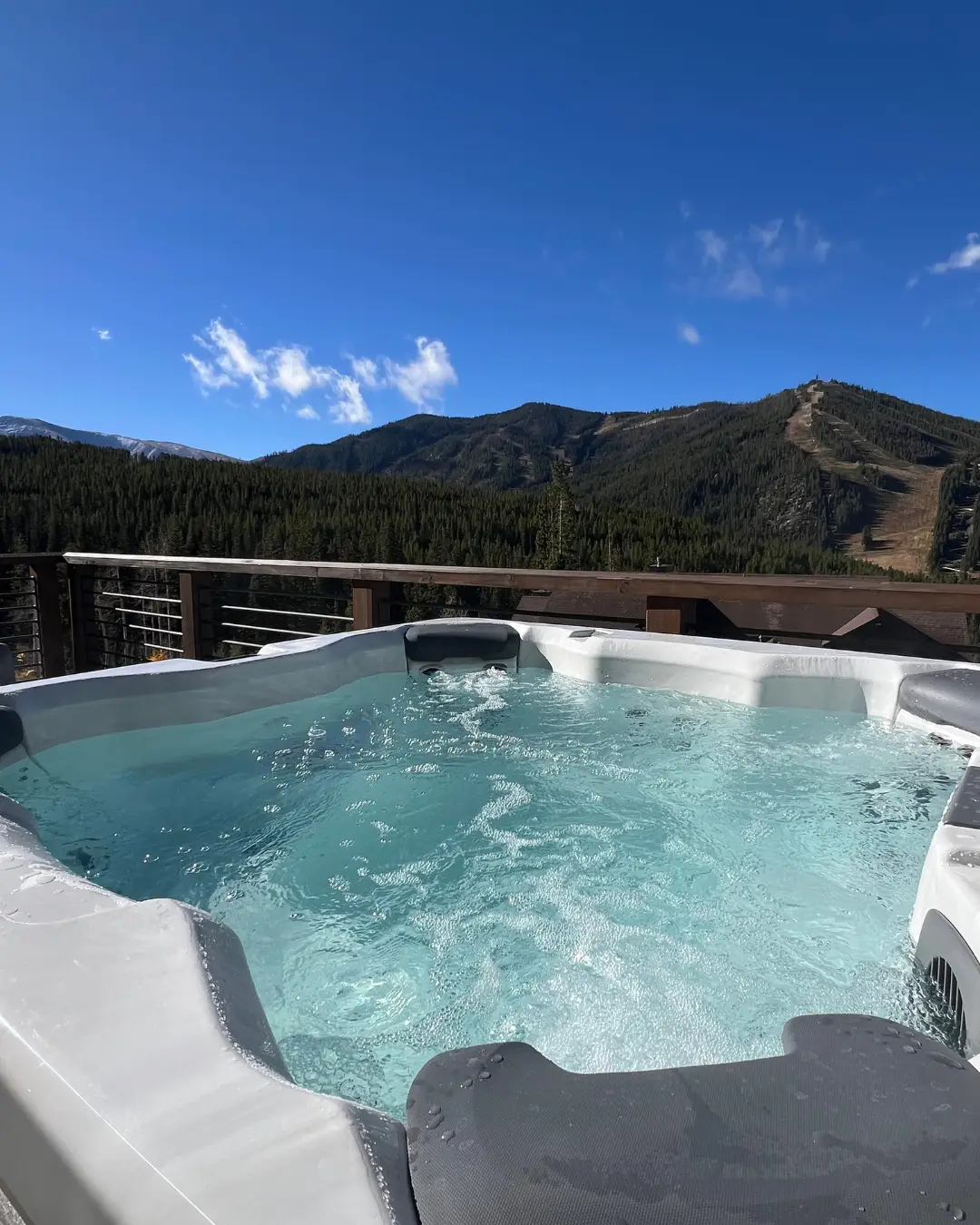 Outdoor hot tub on a deck overlooks forested mountain slopes under a clear blue sky.