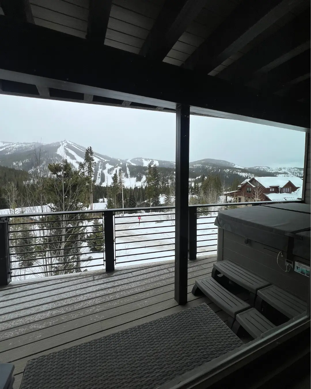 Snow-covered mountain slopes and forest are viewed from a covered deck with a hot tub and railing.