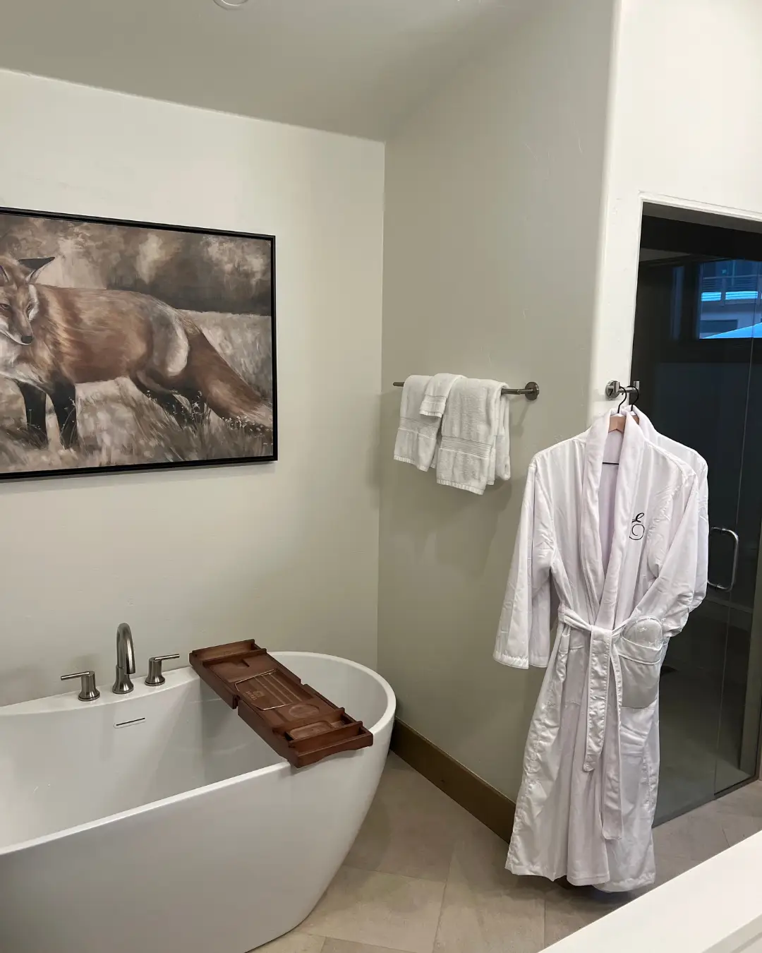 A bathroom with a freestanding tub, wooden bath tray, white robe, towels, glass shower, and fox artwork.