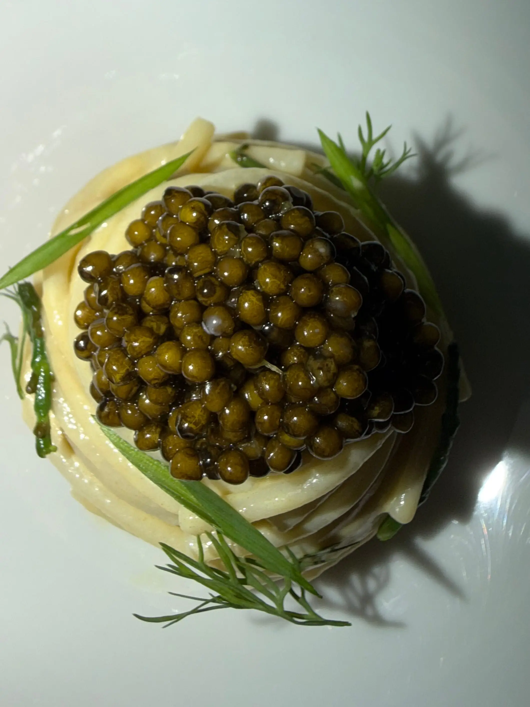 A plated pasta nest topped with caviar and green herbs sits in a pale white bowl.