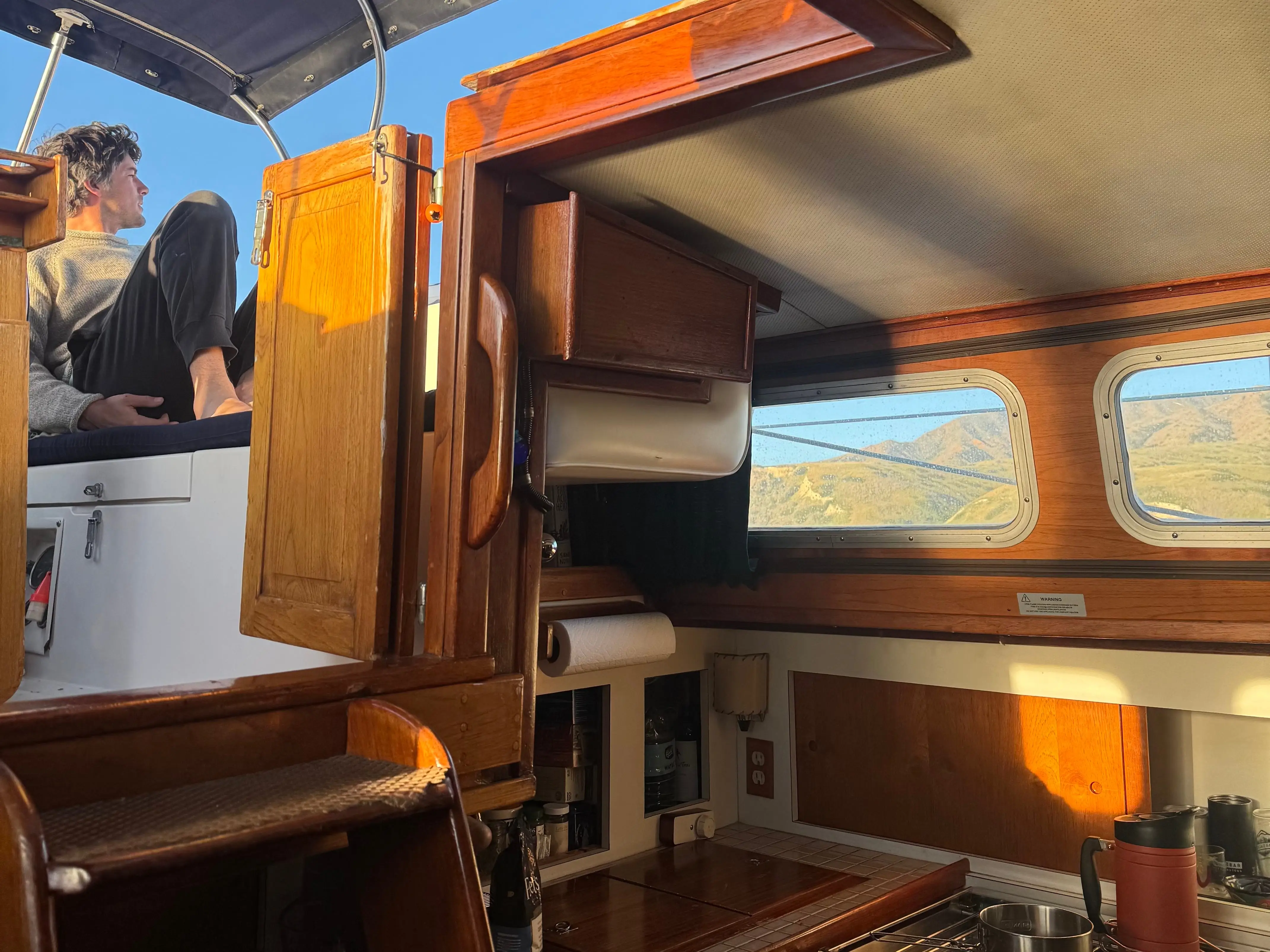 Sunlit wooden boat cabin with galley fixtures, mountain views through windows, and the writer's husband is seated on the deck.
