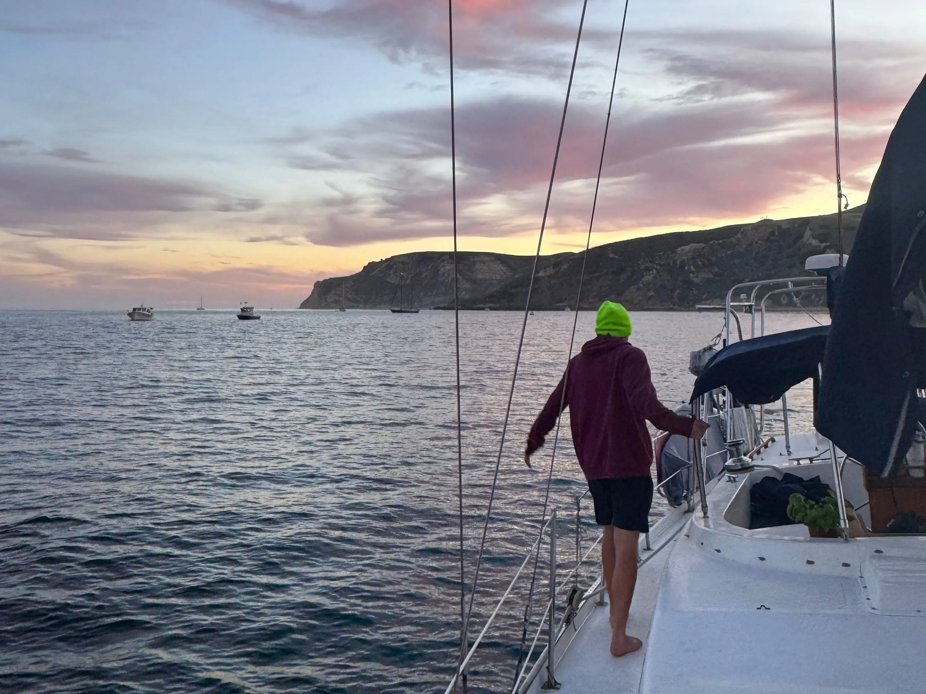 The writer's husband stands on a sailboat deck near a rocky coastline at sunset.
