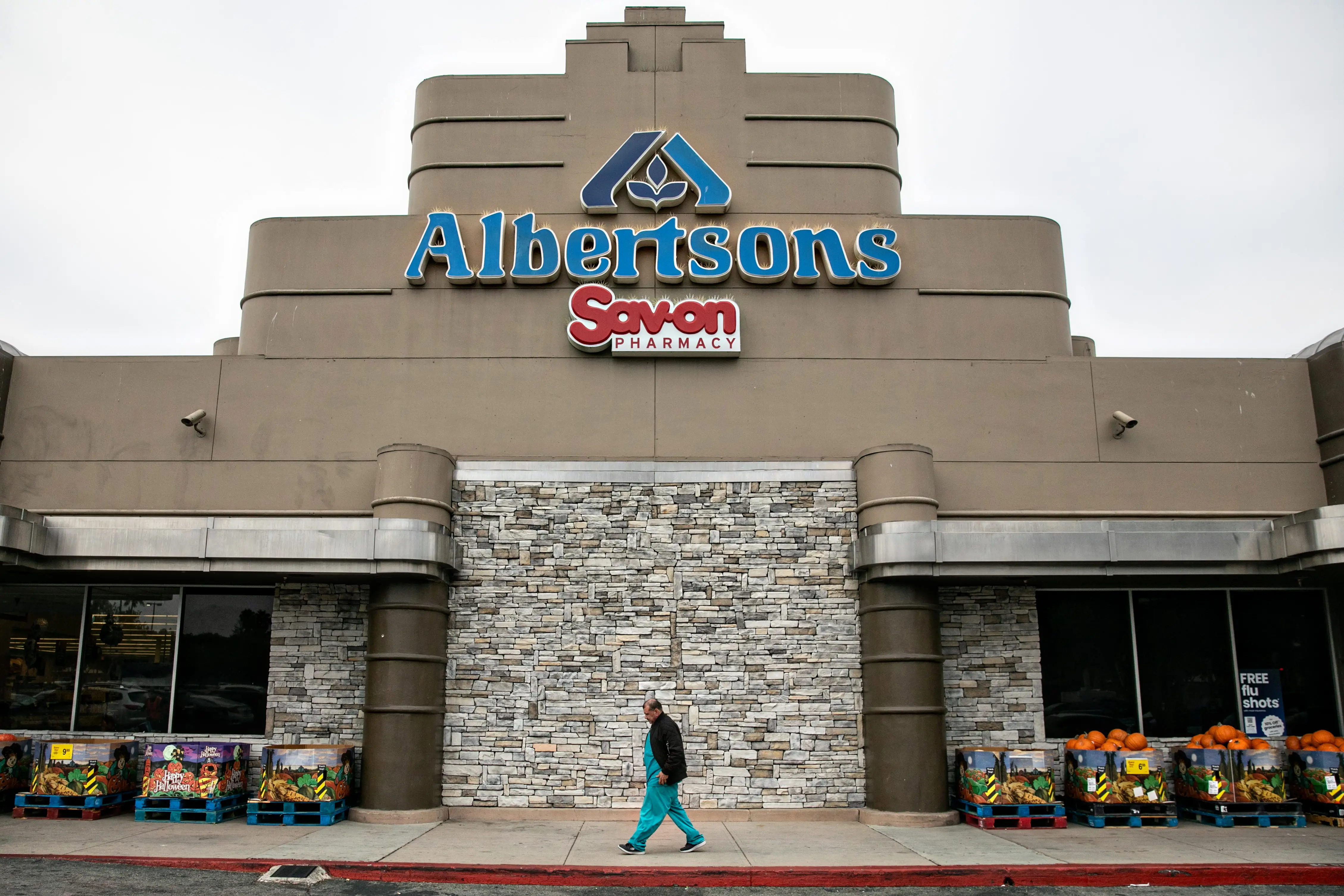 Albertsons storefront with Savon PHARMACY signage and outdoor produce displays as a person walks past.