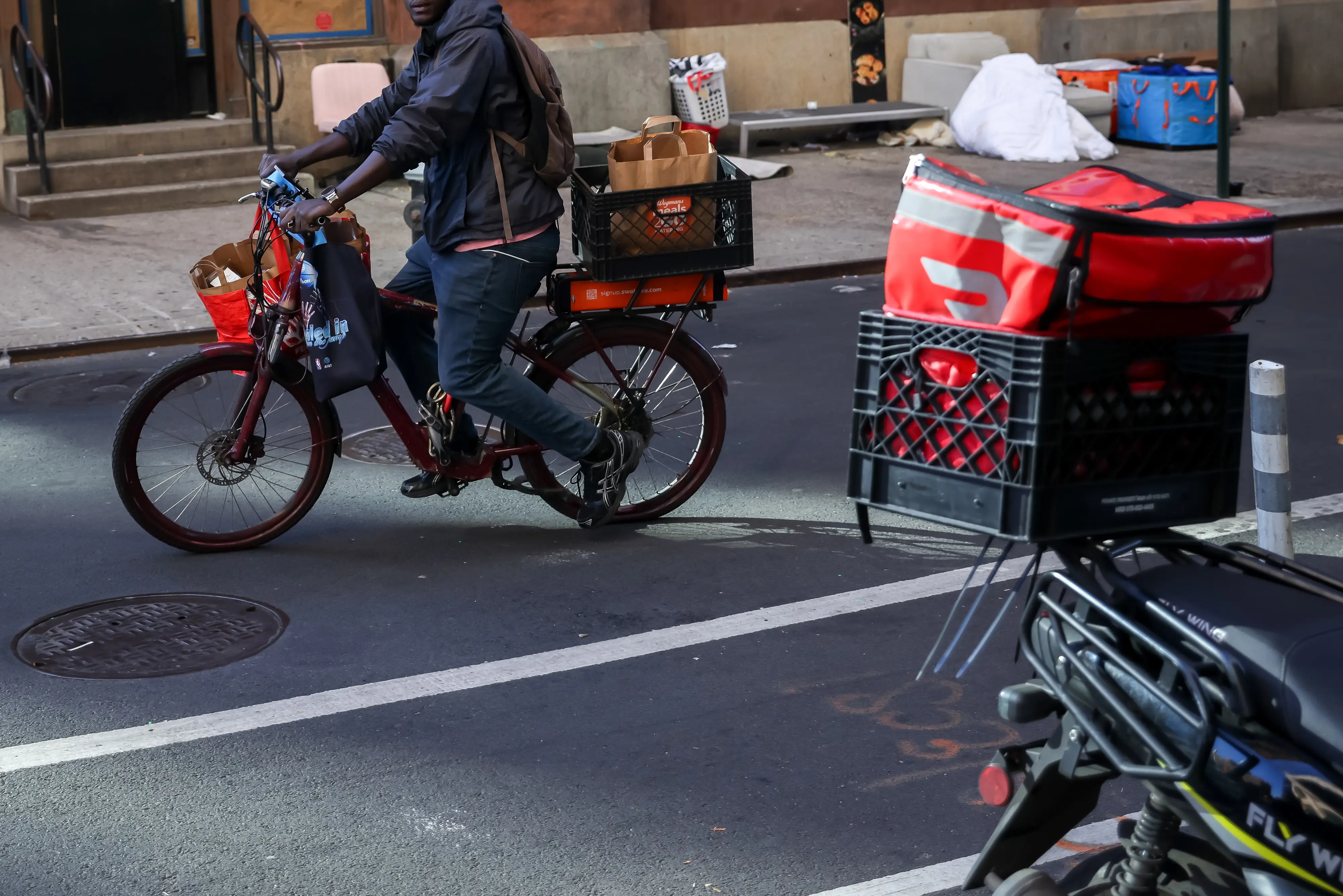 A delivery cyclist rides past a parked bike with a red DoorDash bag in a city bike lane.