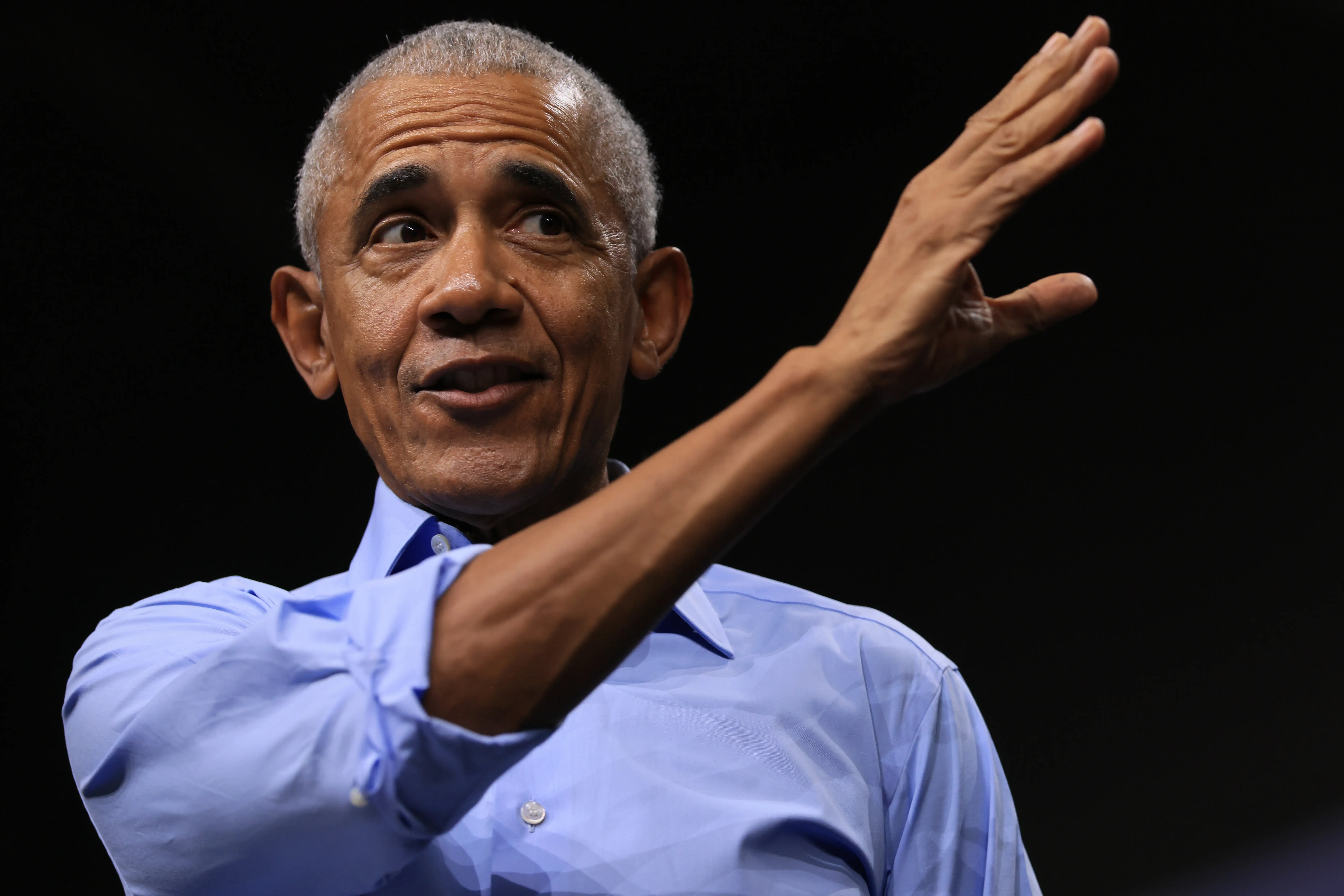 Barack Obama gestures with a raised hand while wearing a light blue shirt against a dark background.