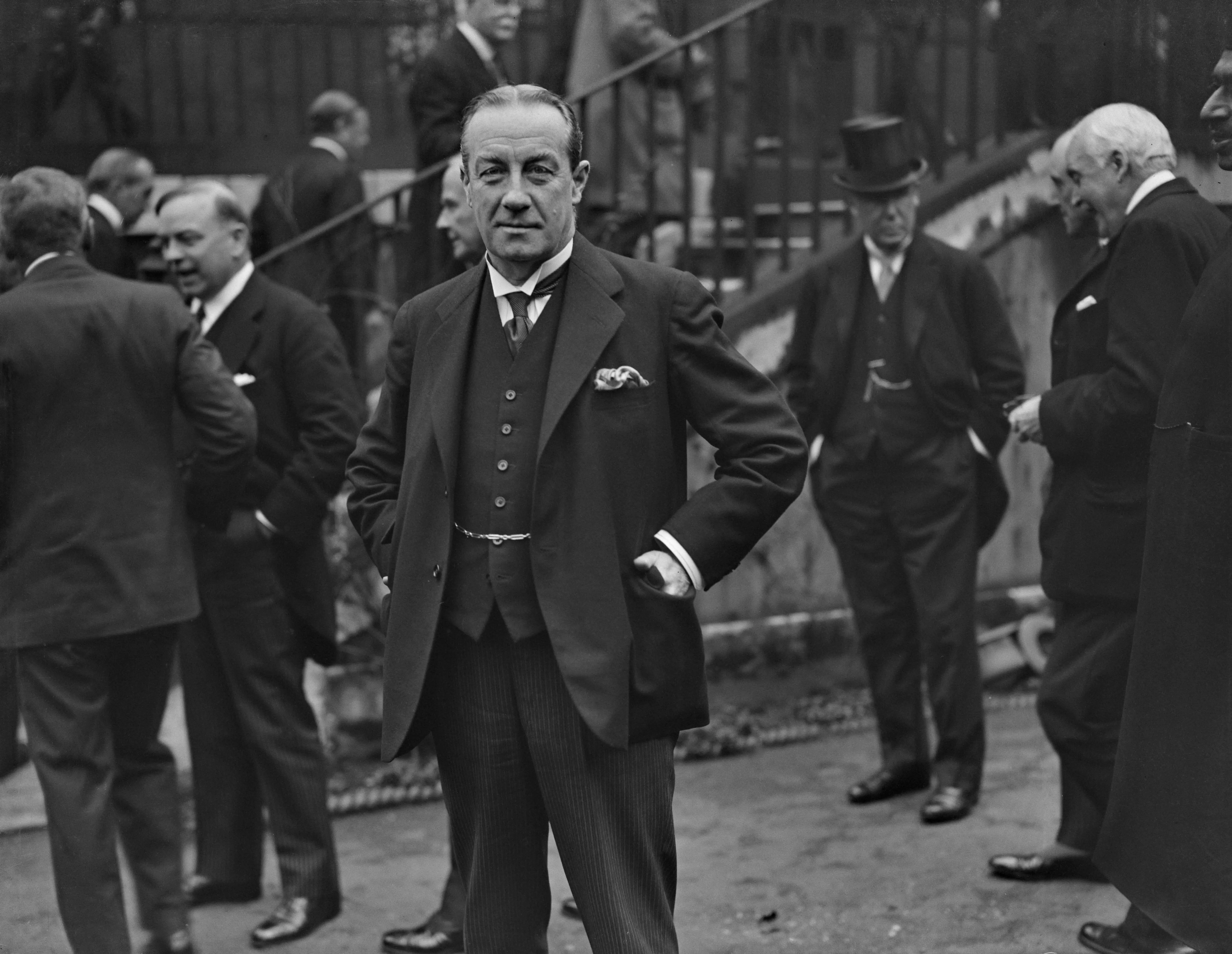 A formally dressed person stands among others in suits outside a building in a black-and-white street scene.