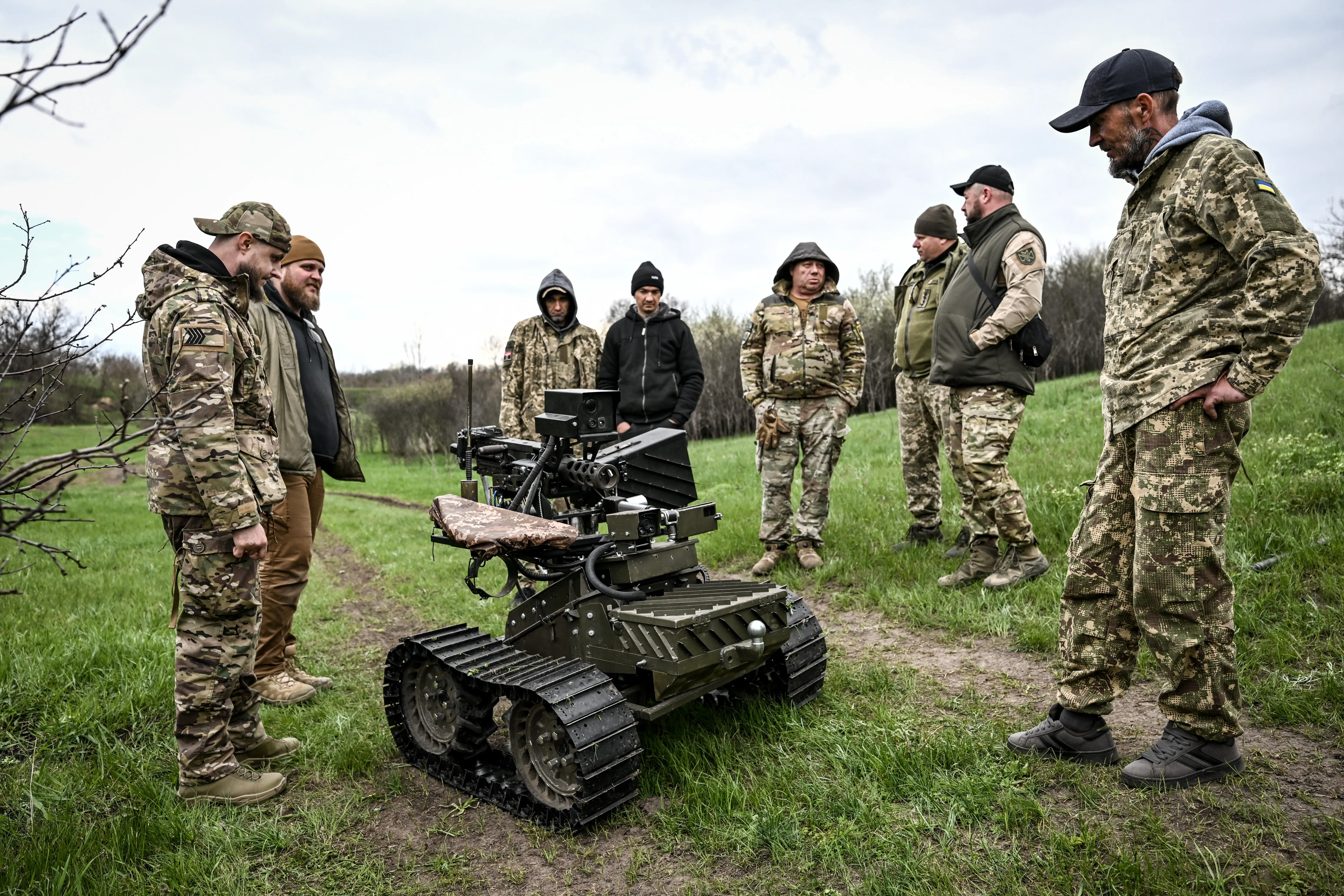 Camouflaged military personnel stand around a tracked ground robot on a grassy path.