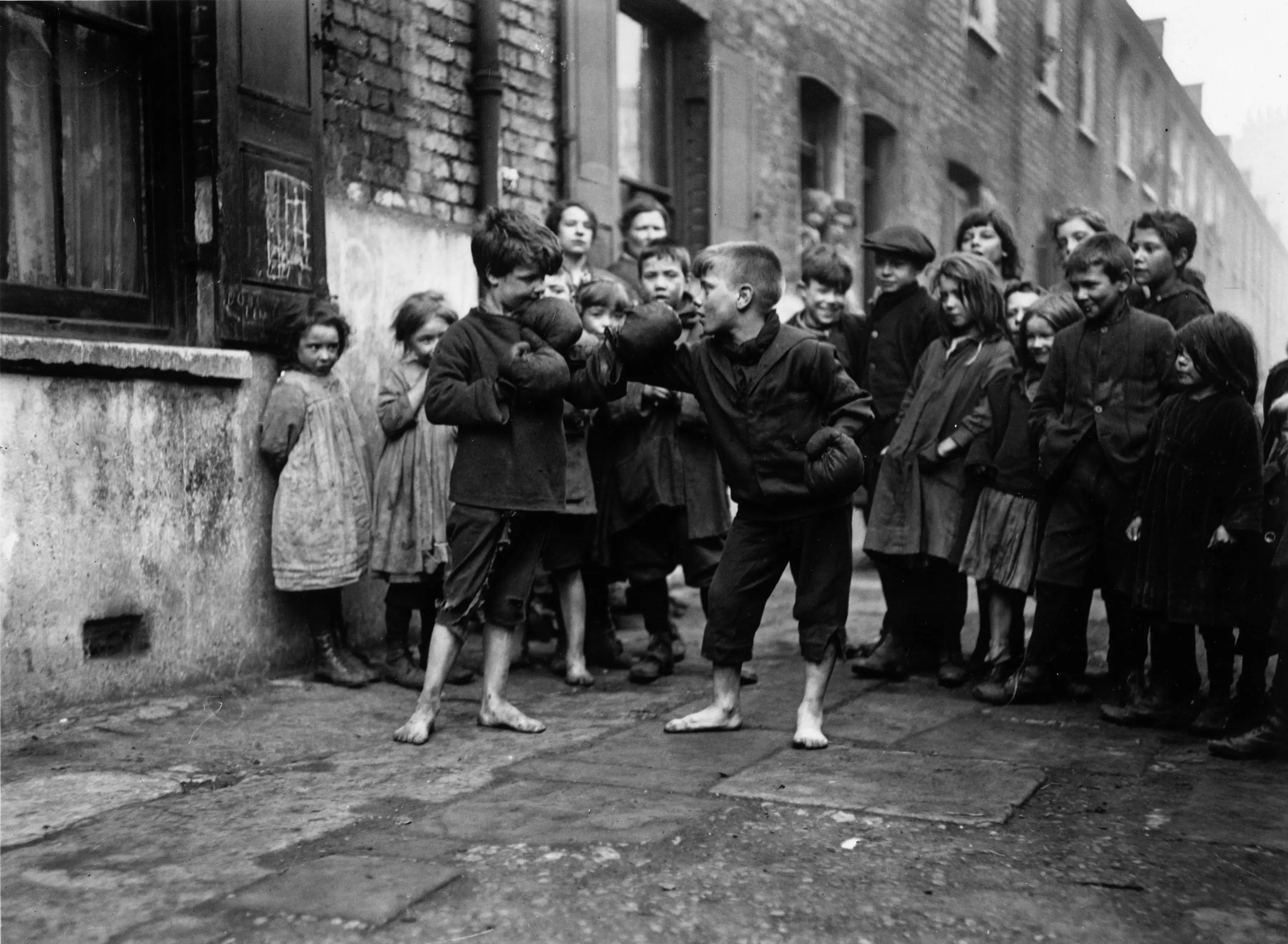 Barefoot people gather in a narrow brick alley as two central figures square off.