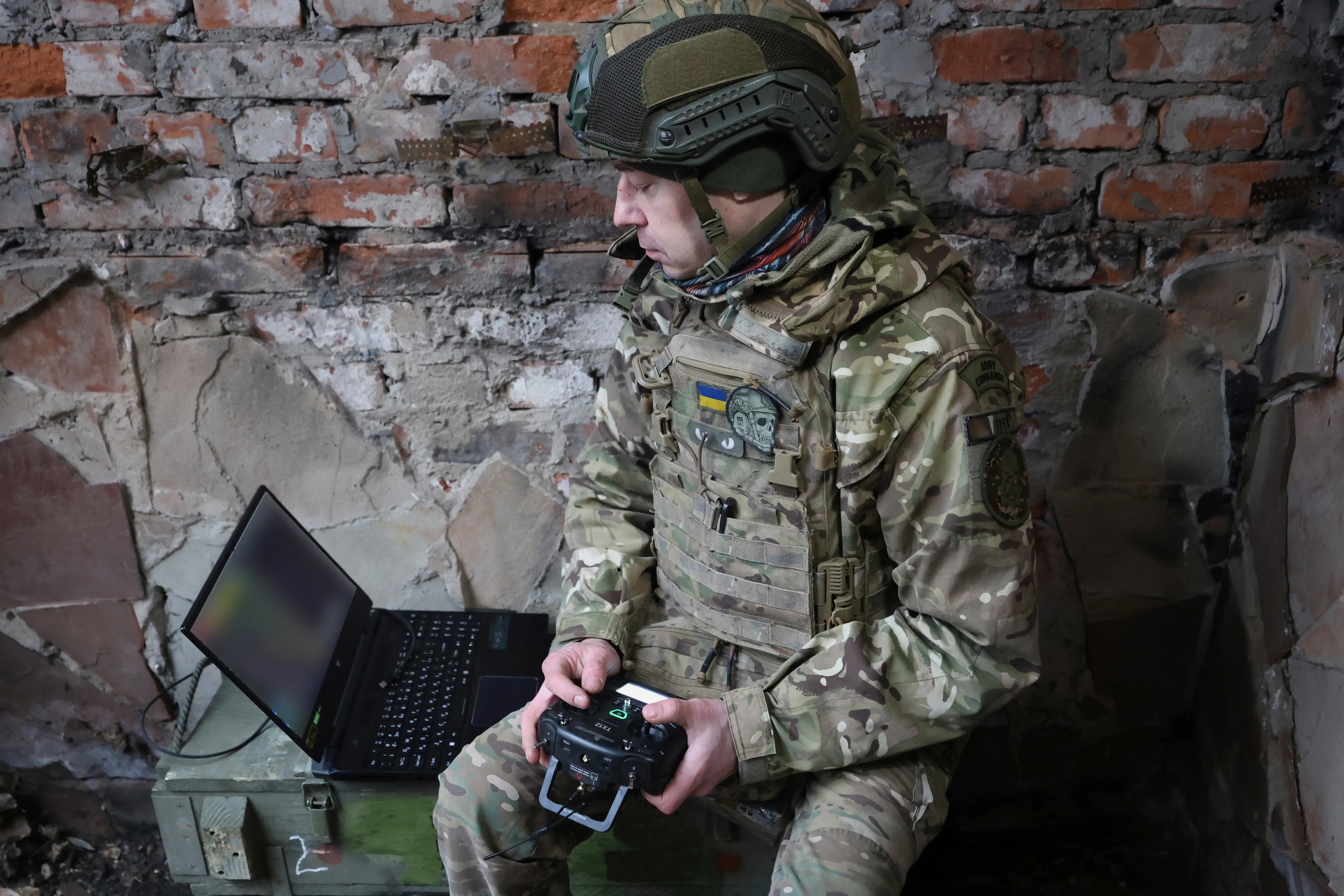 Camouflaged service member with a Ukraine flag patch operates a controller beside a laptop in a damaged brick room.