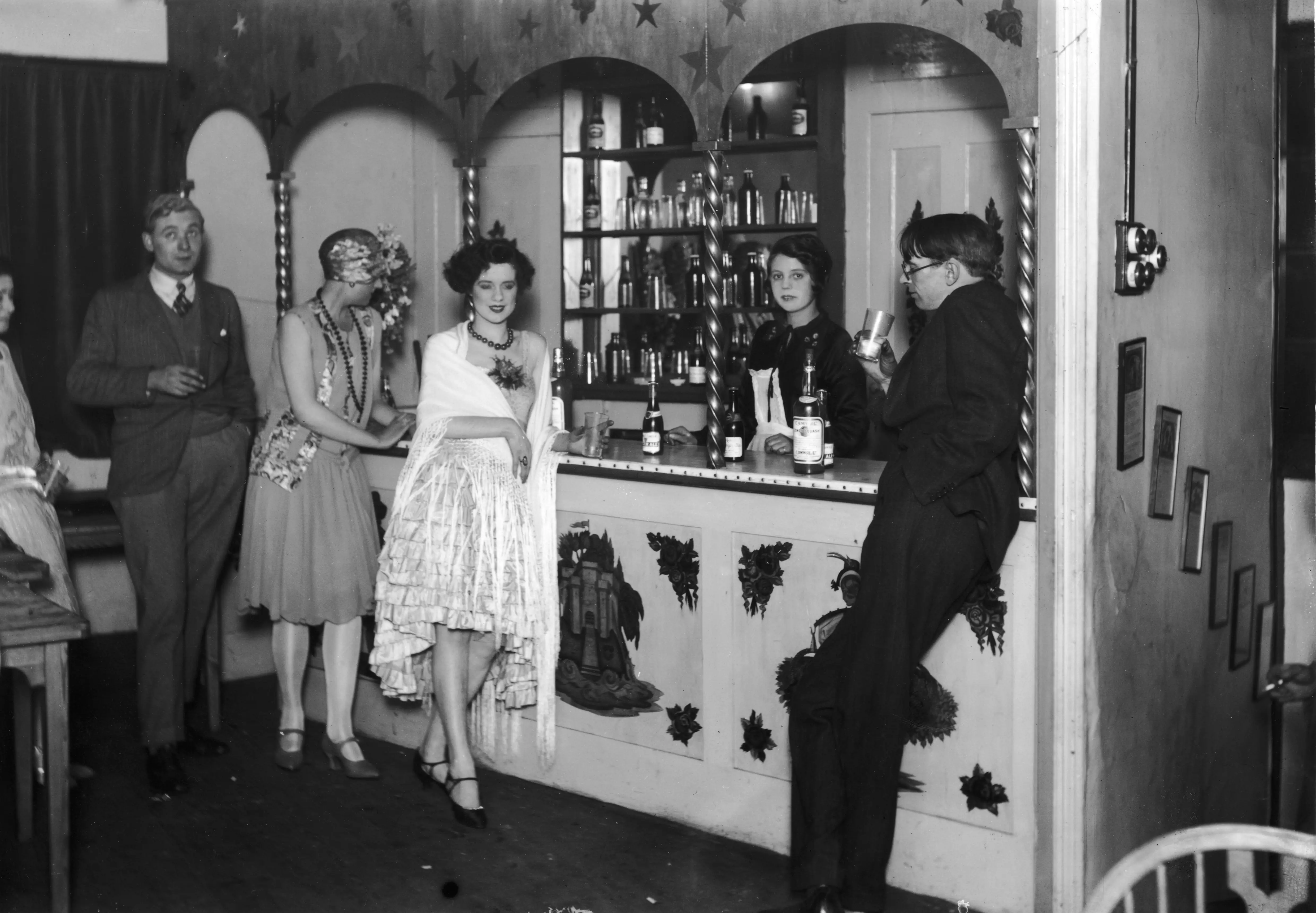 Costumed guests stand around a decorated bar during a vintage indoor party in black and white.