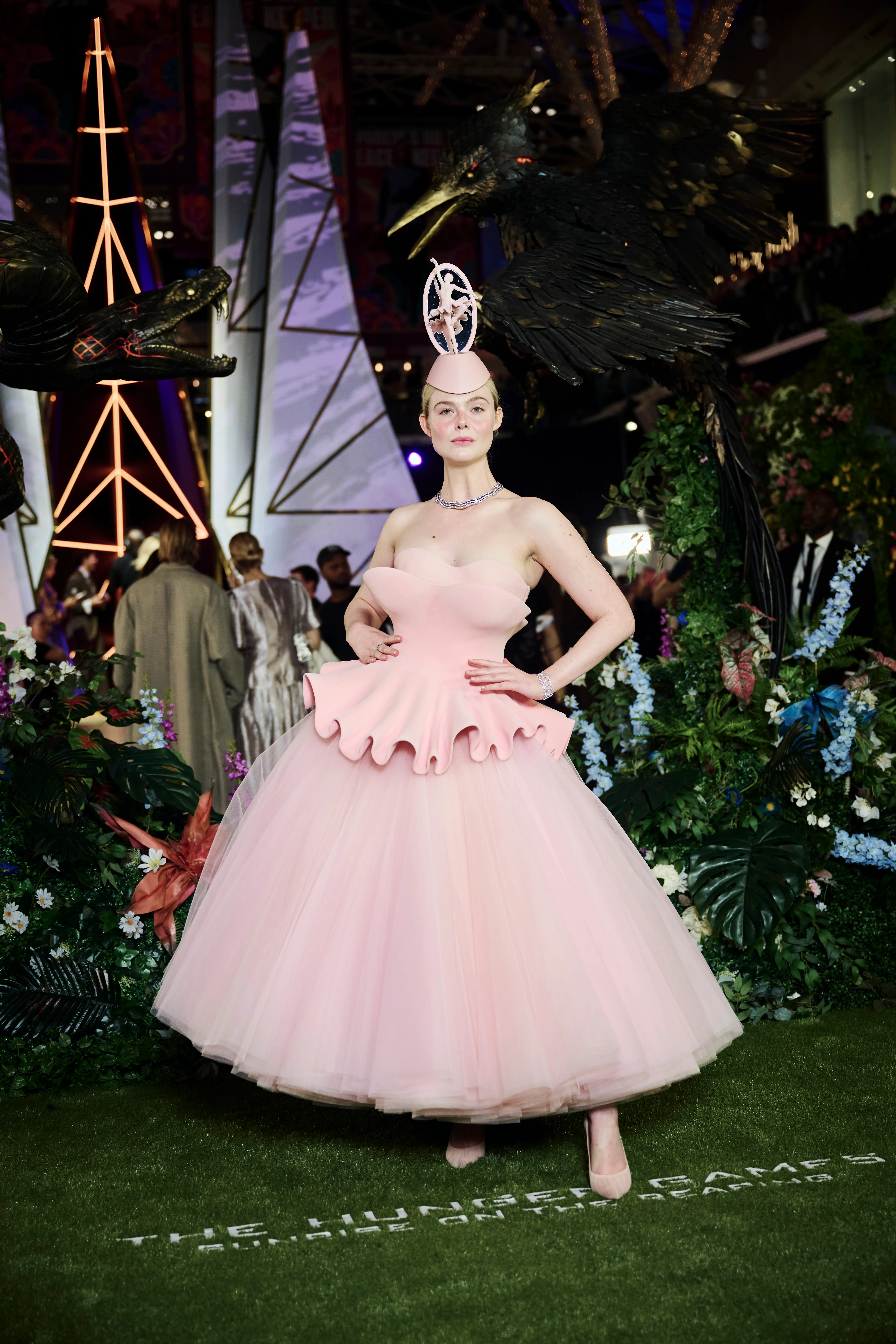 Elle Fanning poses in a pink tulle gown amid floral decor and bird sculptures at a The Hunger Games event.