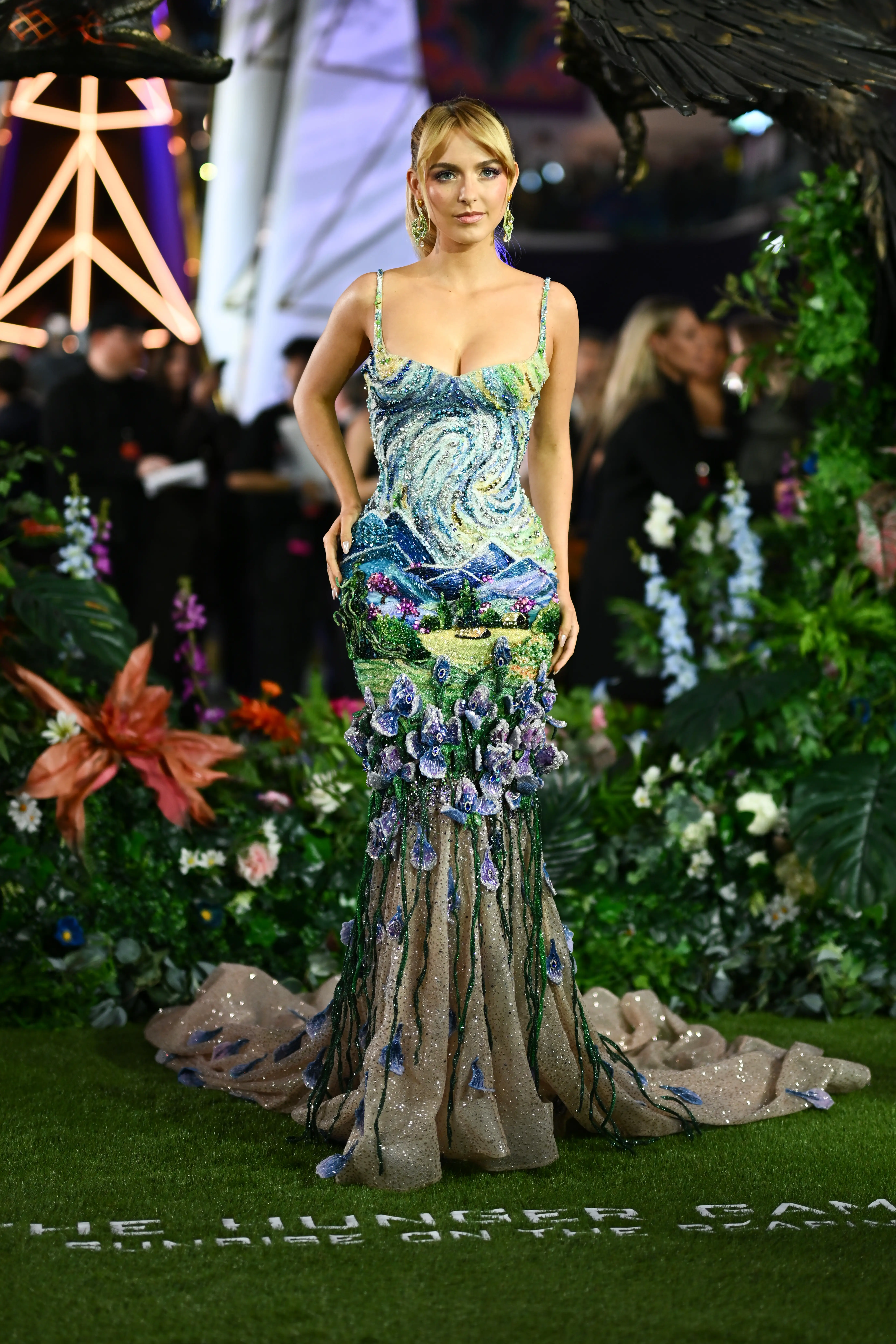 McKenna Grace in an ornate beaded floral gown stands on a green carpet amid lush garden décor.