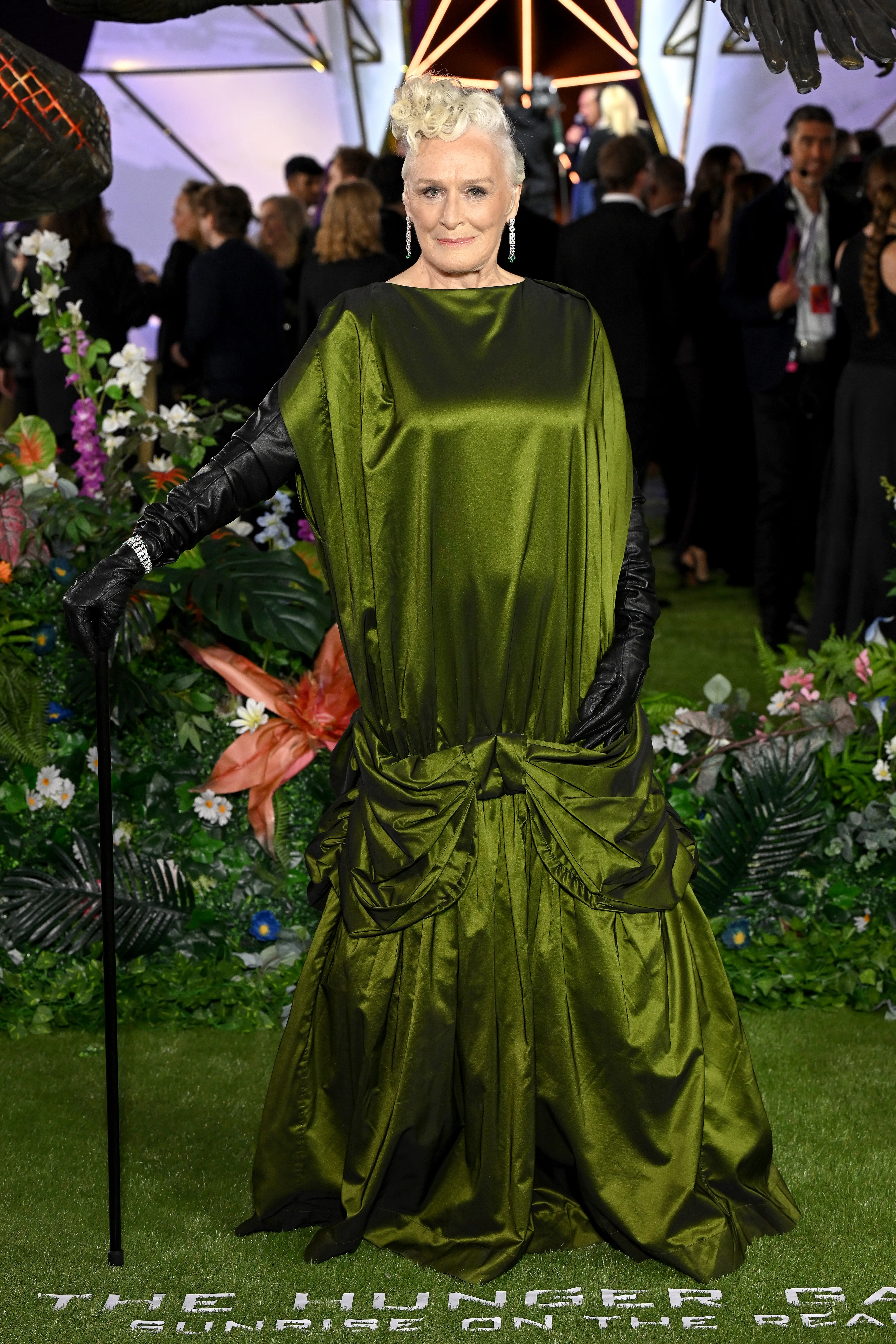 Glenn Close poses in a green draped outfit on a floral carpet at a Hunger Games event.
