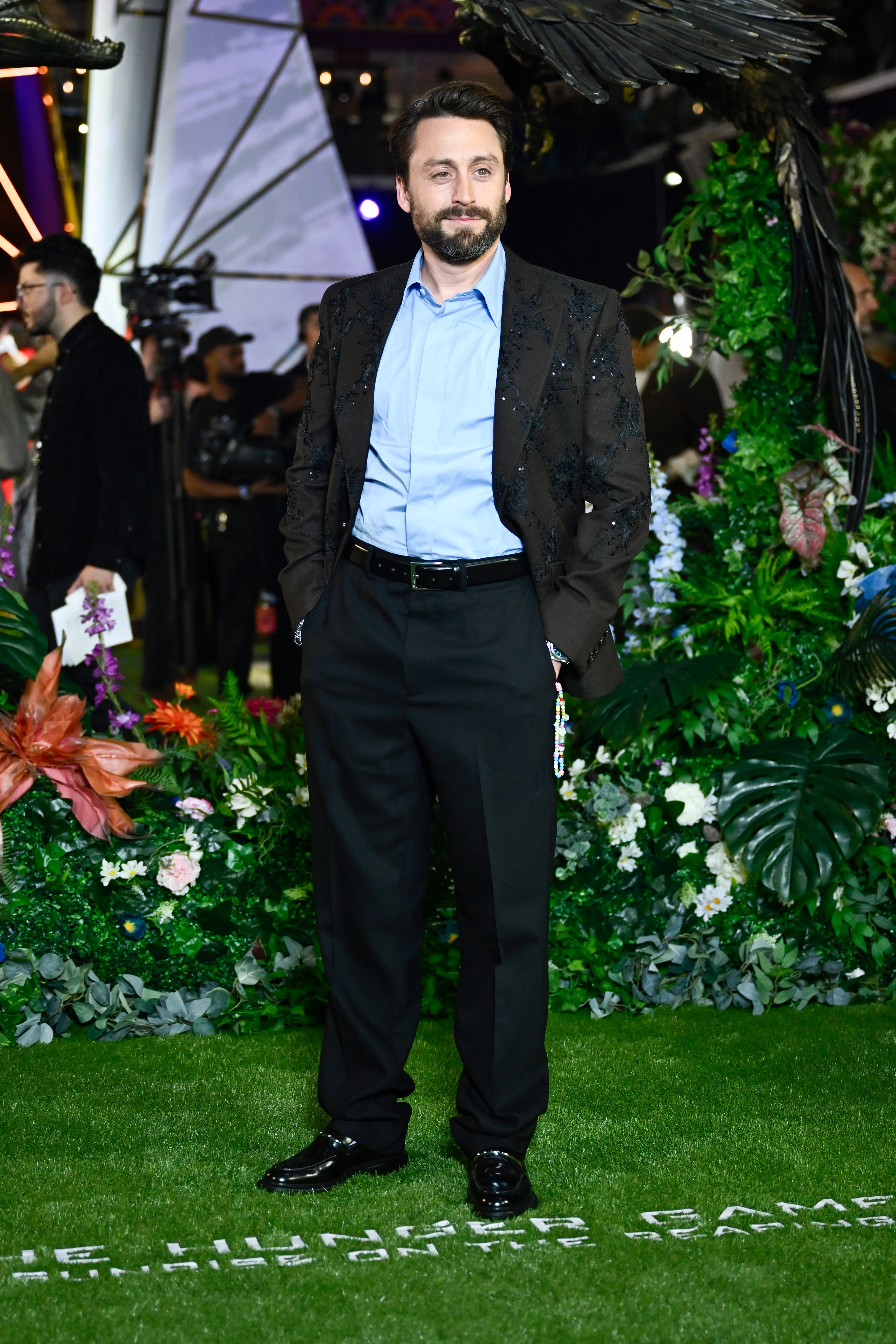 Kieran Culkin poses on a green carpet surrounded by lush plants and event lighting.