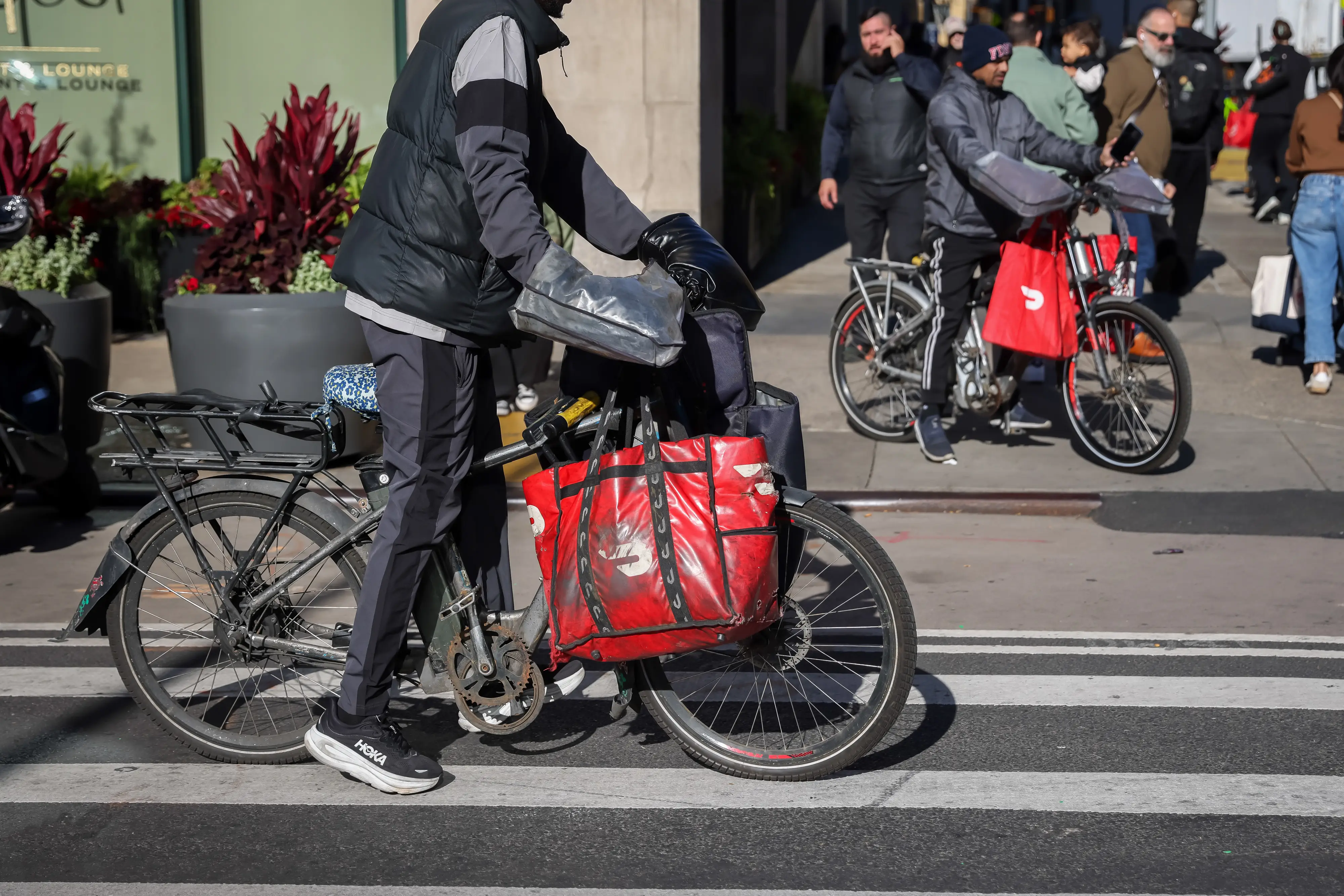 Delivery cyclists with red insulated bags wait with bicycles at a city crosswalk on a sunny street.