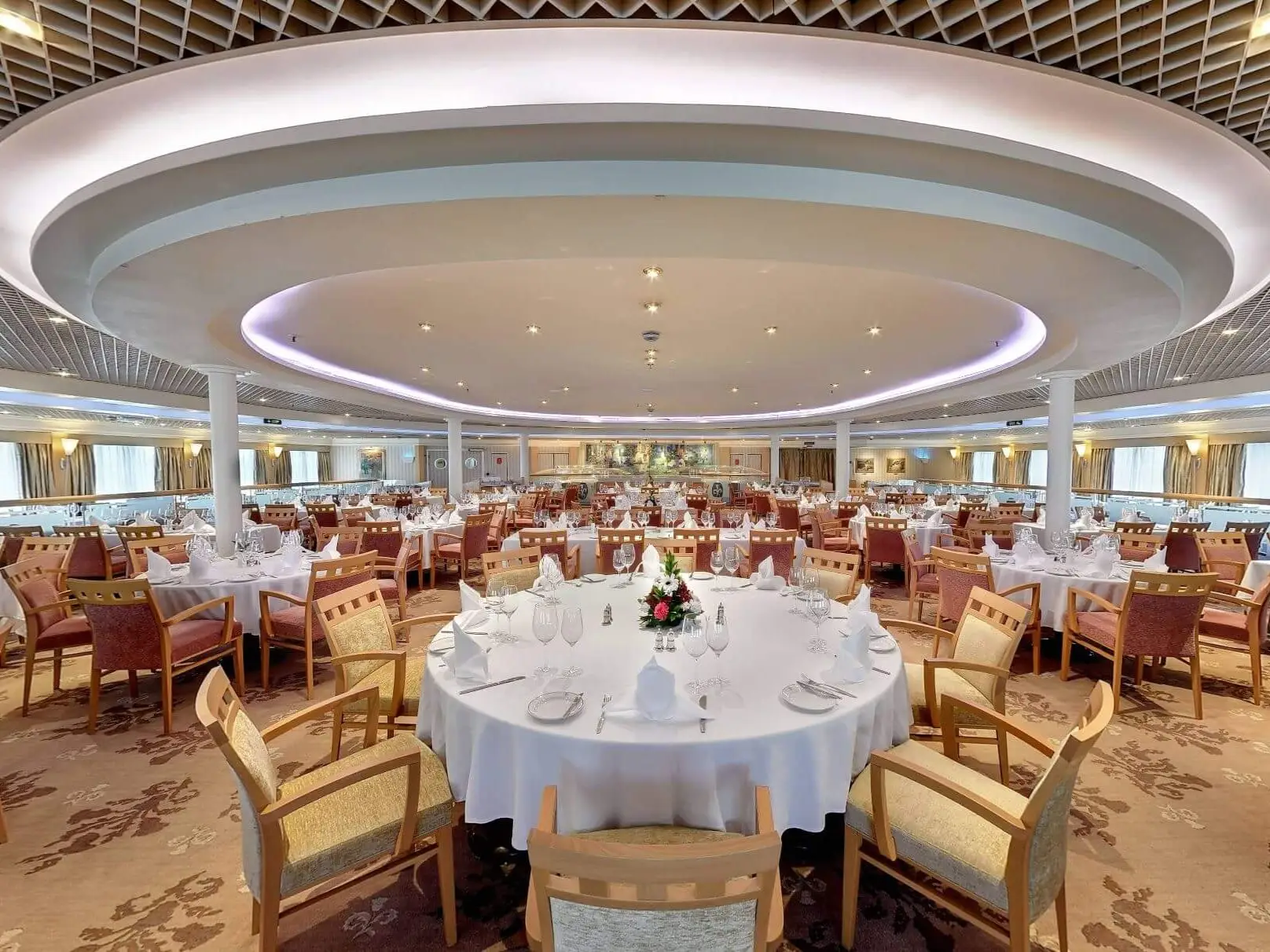 Large formal dining room set with round tables, white linens, glassware, and chairs under a circular ceiling feature.