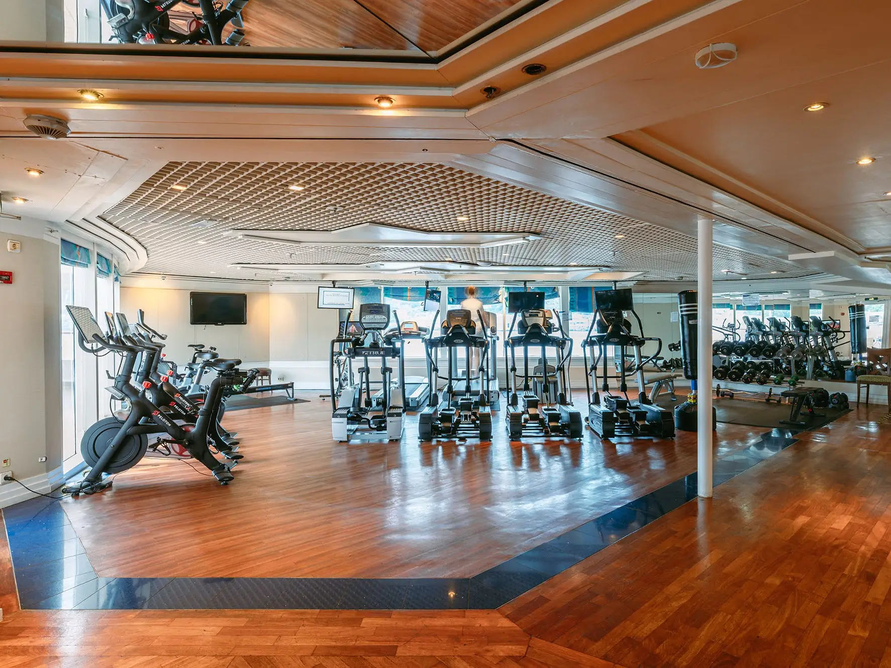 Bright fitness room with treadmills, stationary bikes, weights, mirrors, and one person exercising on a treadmill.