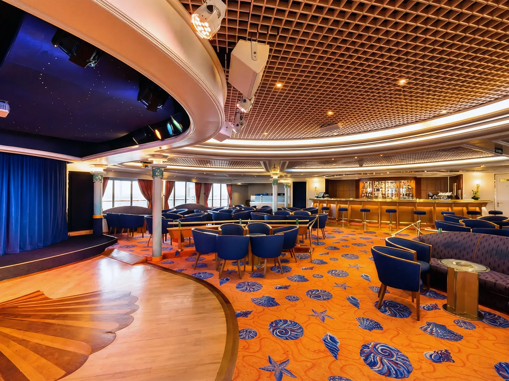 Lounge with a small stage, bar, blue seating, and shell-patterned carpet in a warmly lit interior.