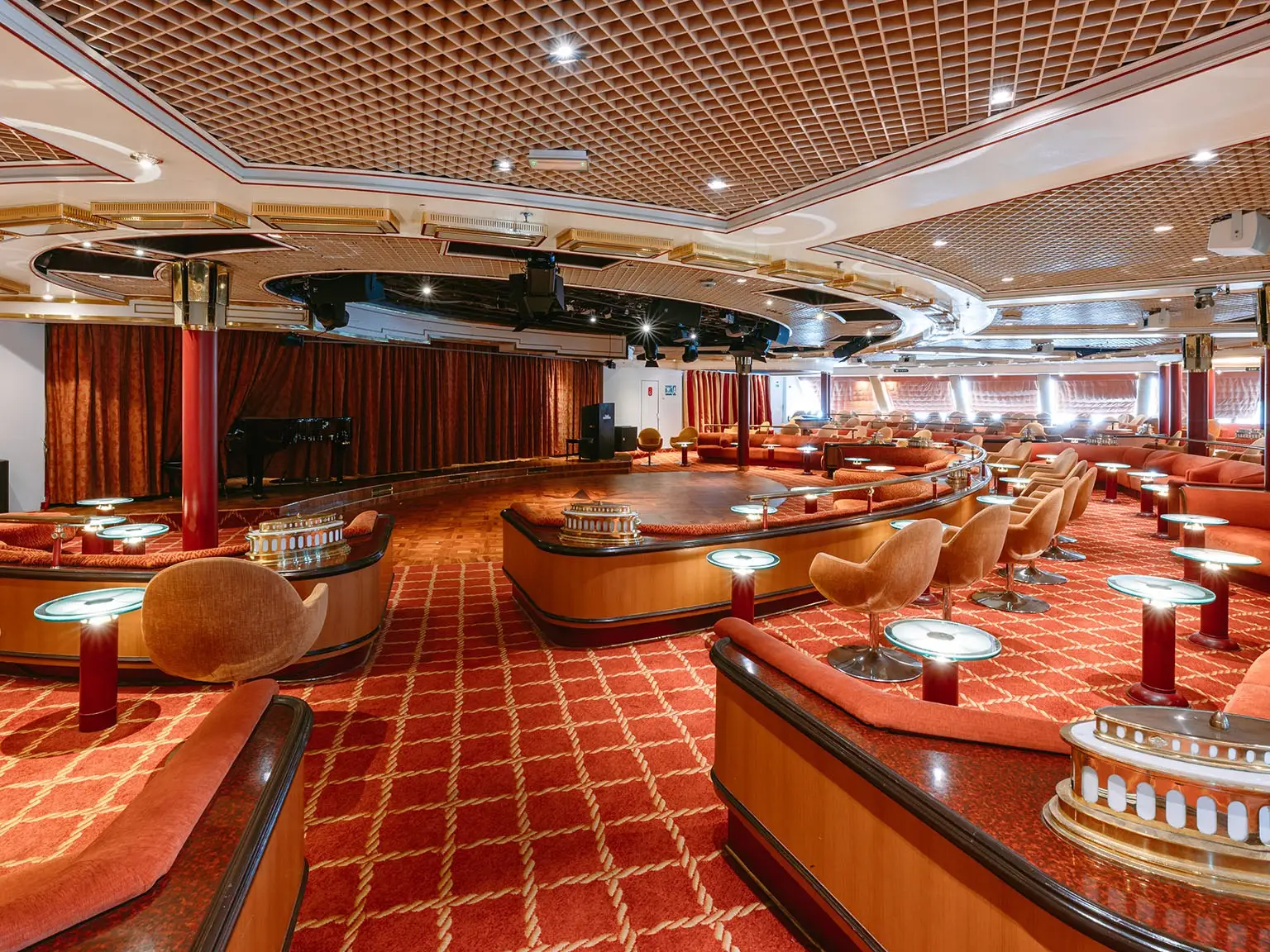 Empty lounge with red seating, cocktail tables, a stage, curtains, and a grand piano under a patterned ceiling.