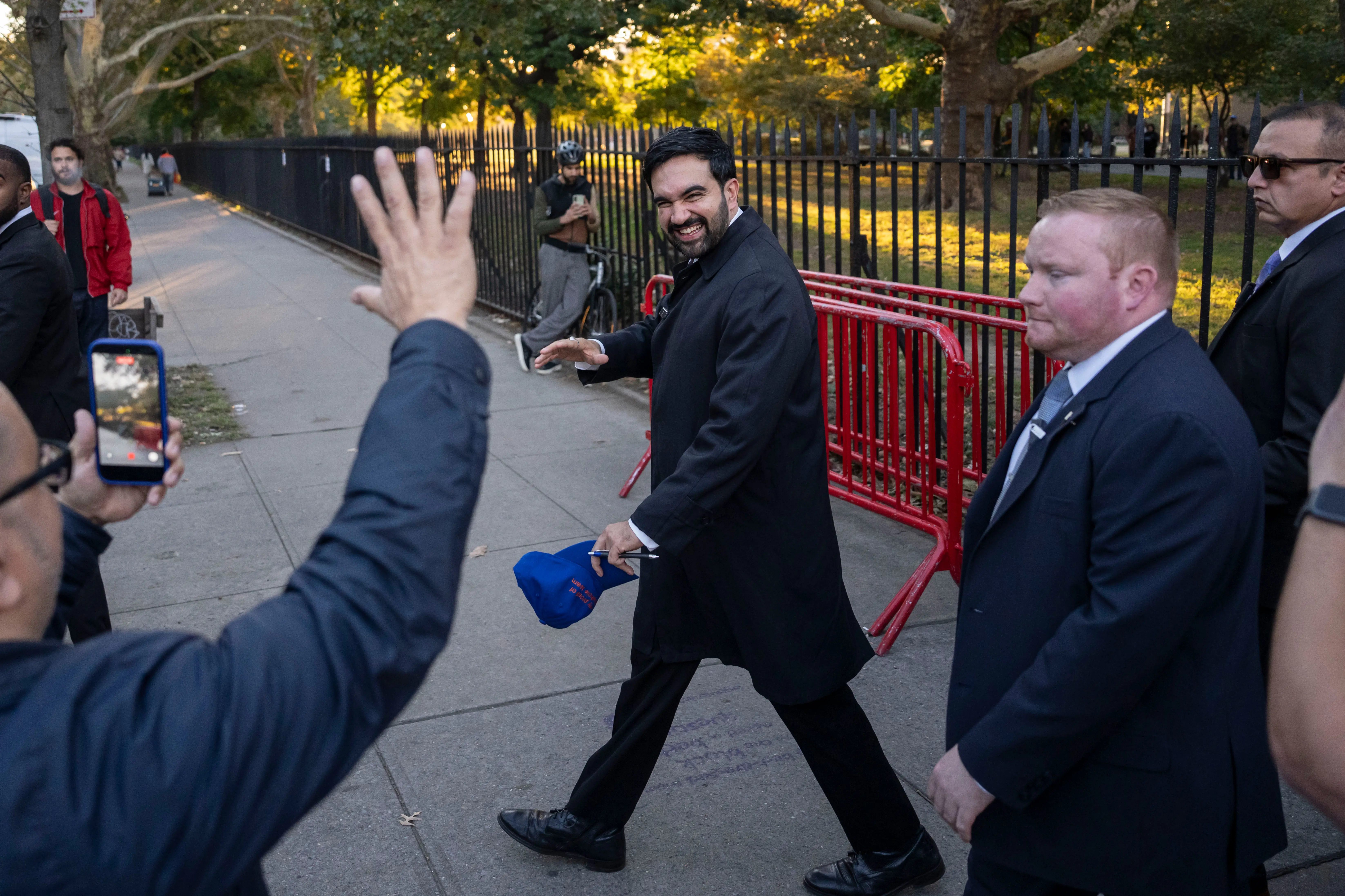 Zohran Mamdani smiles and waves while walking beside red barricades with suited companions on a park-side sidewalk.