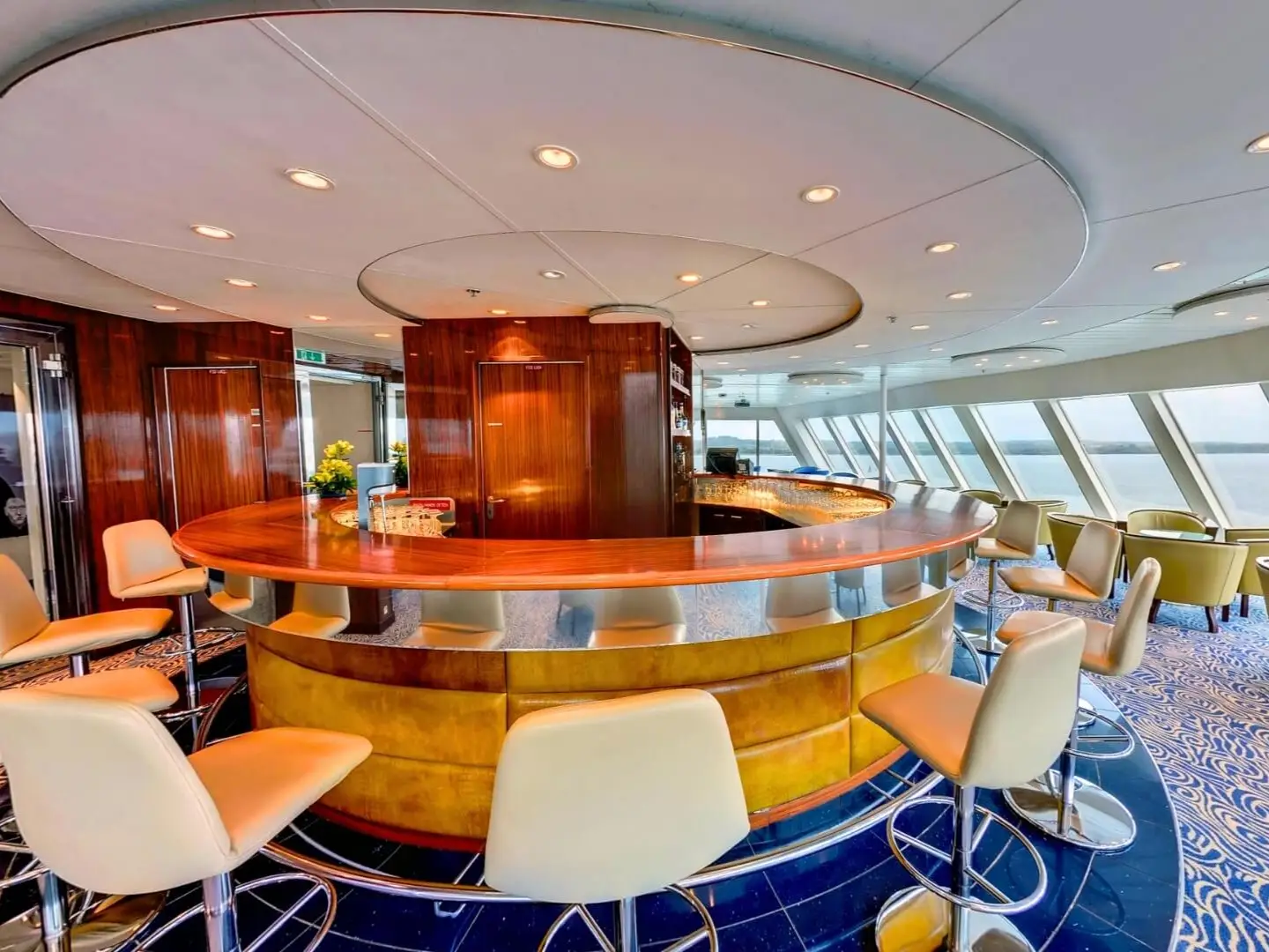 Curved wooden bar with cream stools inside a lounge overlooking water through large slanted windows.