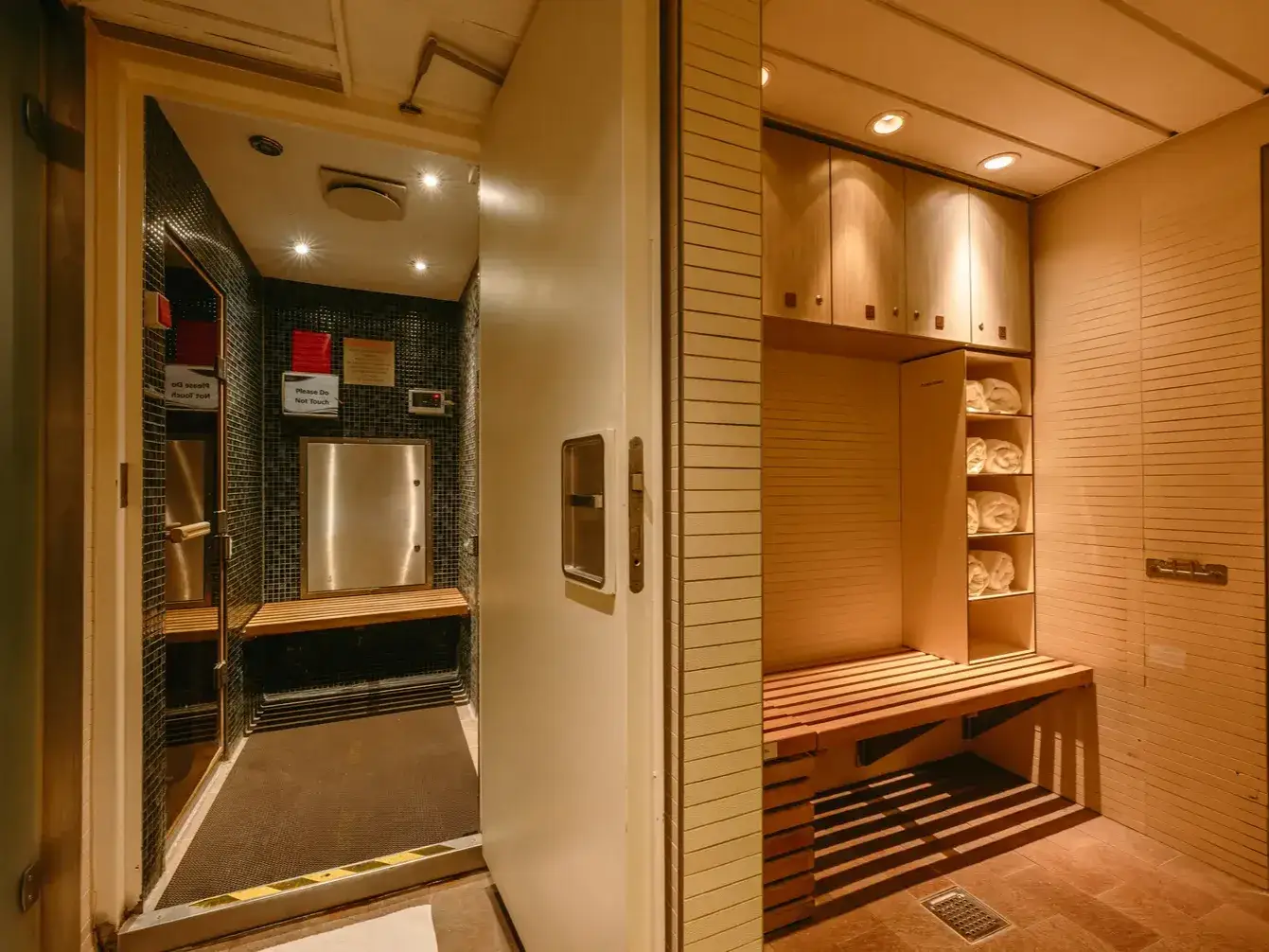 Warmly lit changing area with wooden bench, towels, cabinets, shower room, and tiled walls.