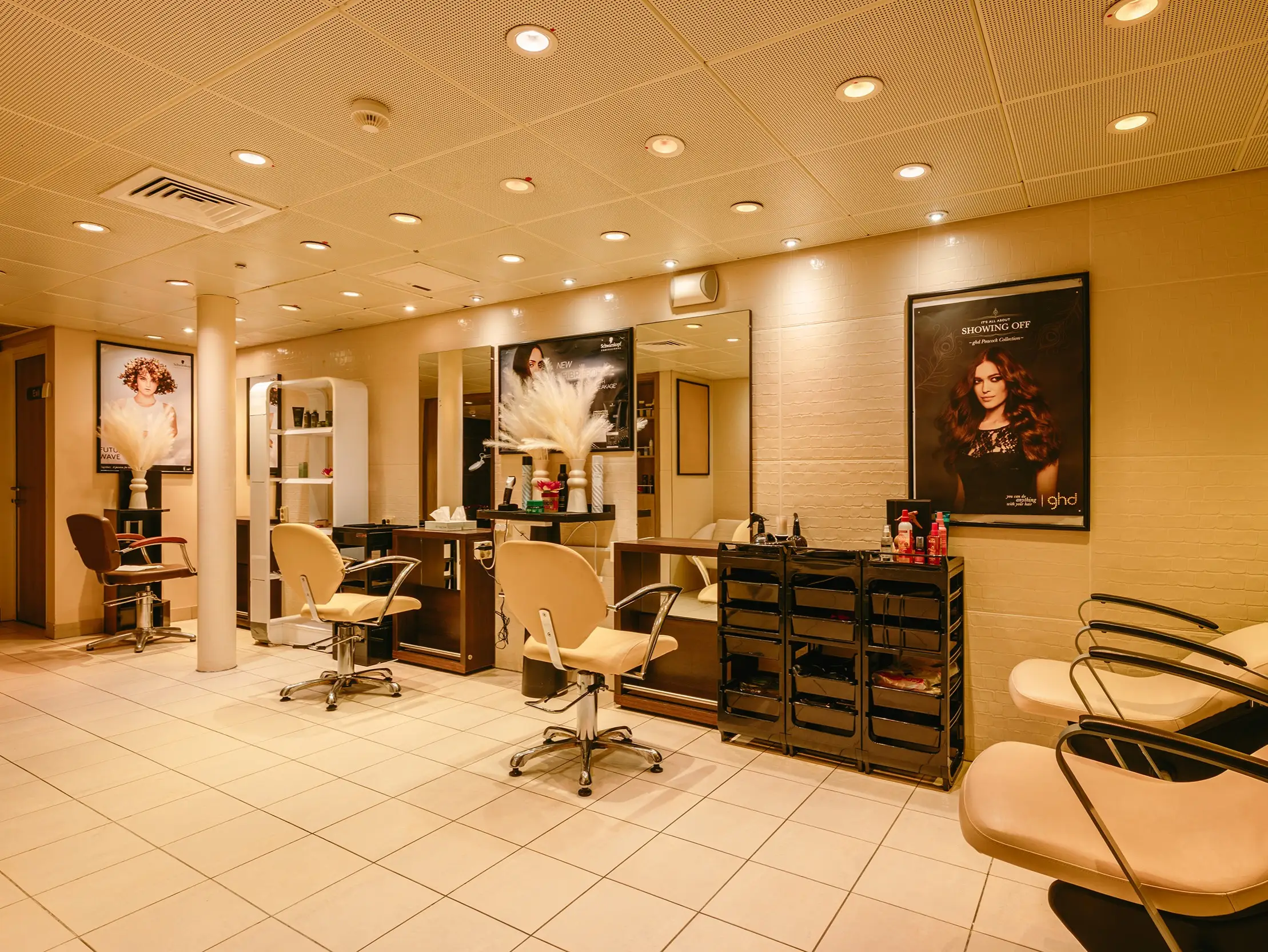 Warmly lit hair salon interior with styling chairs, mirrors, product carts, posters, and tiled flooring.