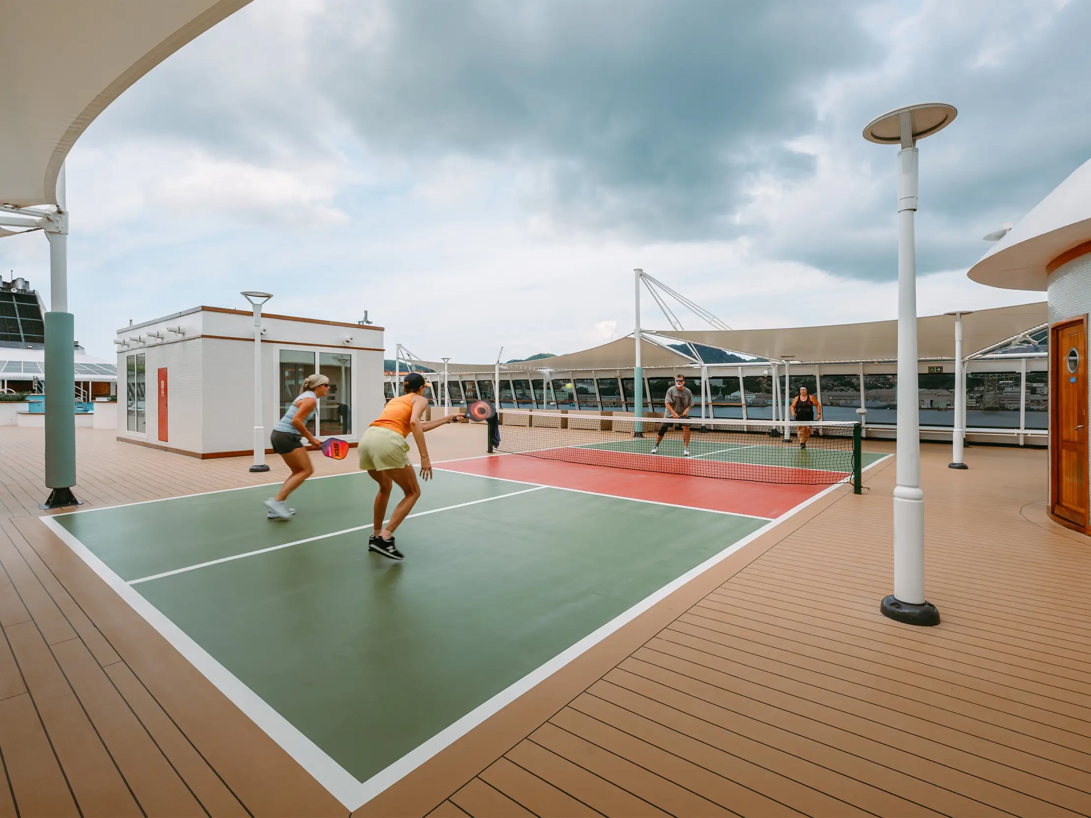 Players rally on an outdoor pickleball court on a ship deck under cloudy skies.