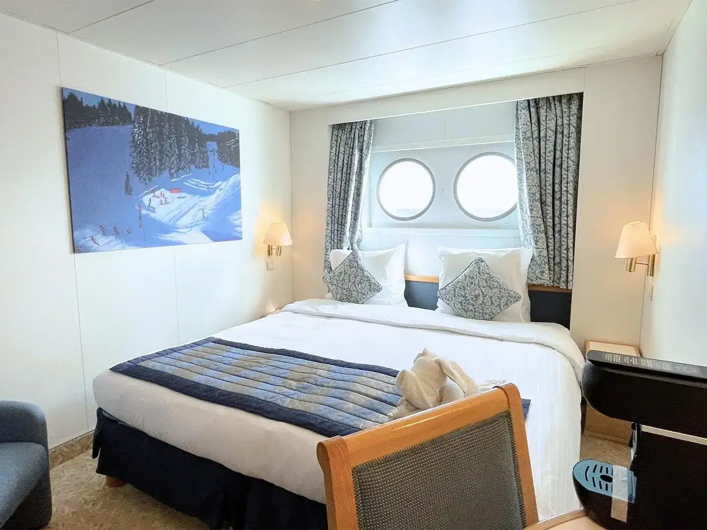 Cruise ship cabin with a double bed, two round windows, wall lamps, curtains, and ski slope artwork.