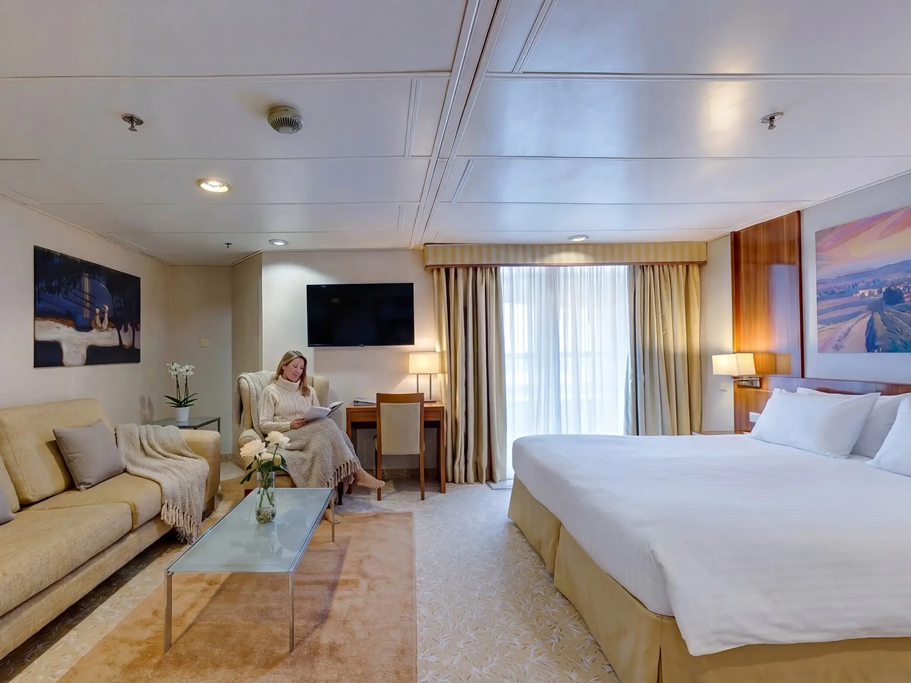 Spacious hotel-style cabin with a large bed, sofa, desk, wall art, and a person seated in an armchair reading.