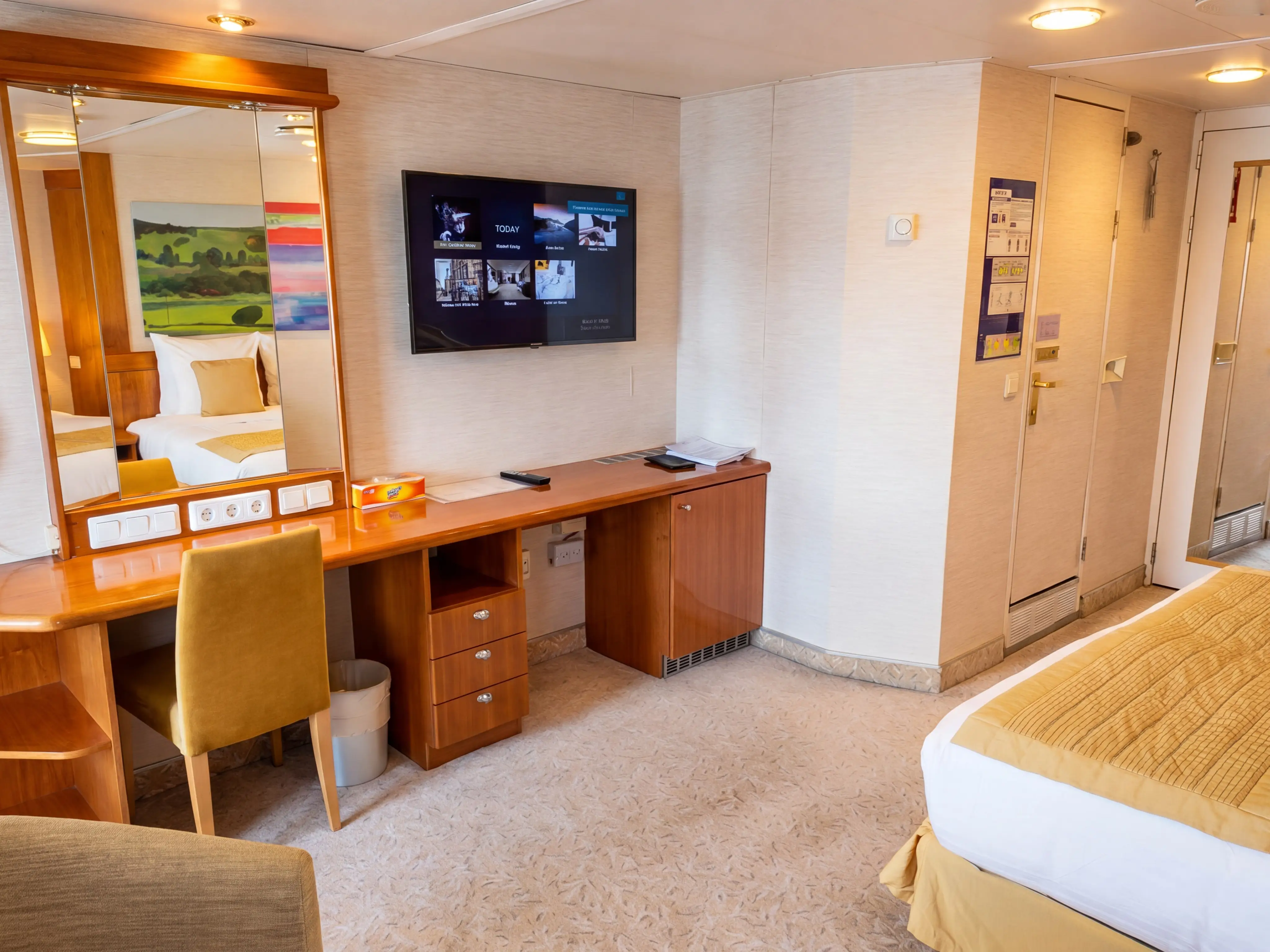 Compact guest room interior with bed, desk, mirror, wall-mounted TV, and door in warm beige decor.