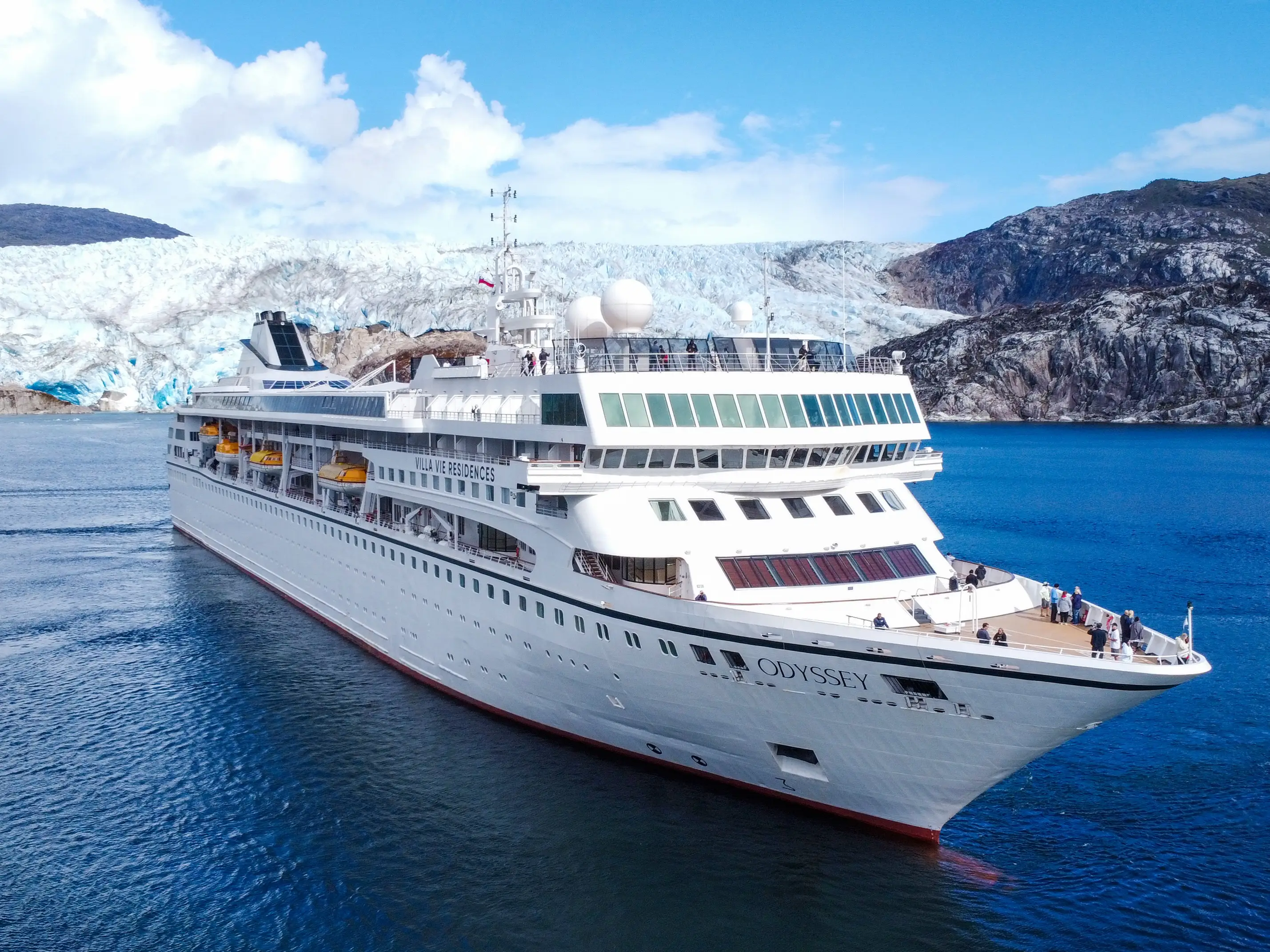 White cruise ship ODYSSEY sails through blue water in front of a glacier and rocky mountains.