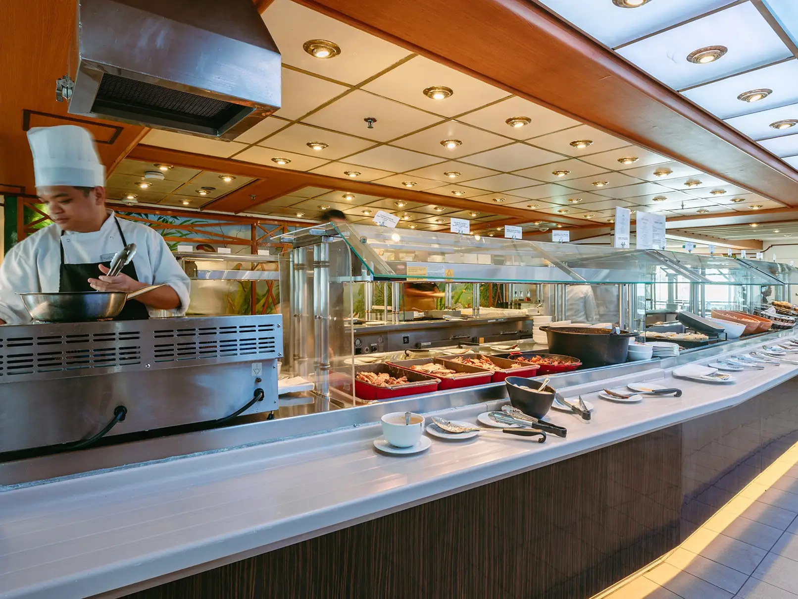 Chef works at a buffet station beside a long restaurant serving counter with food trays and plates.