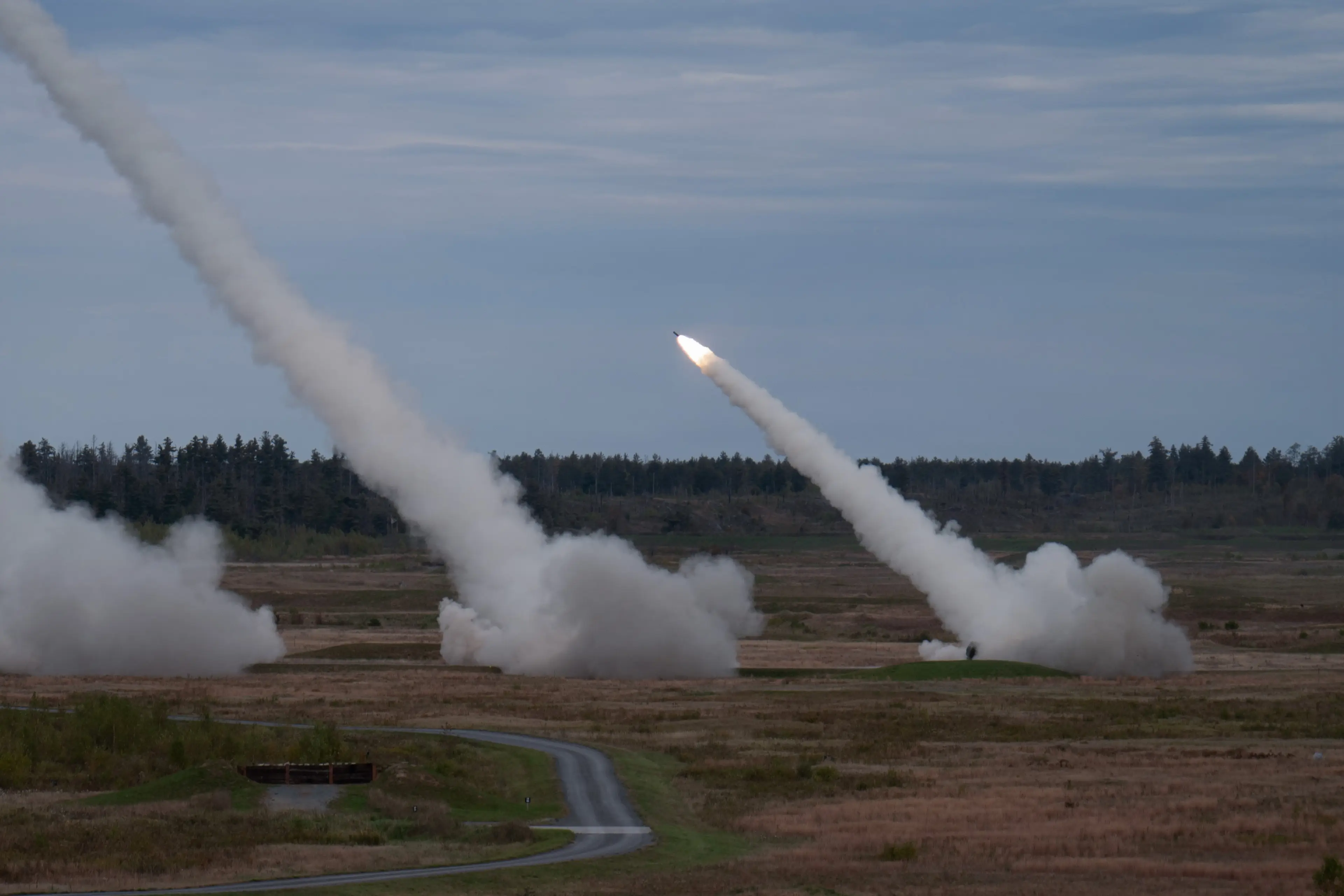 Multiple High Mobility Artillery Rocket Systems fire in a forest terrain with an overcast sky in the background.