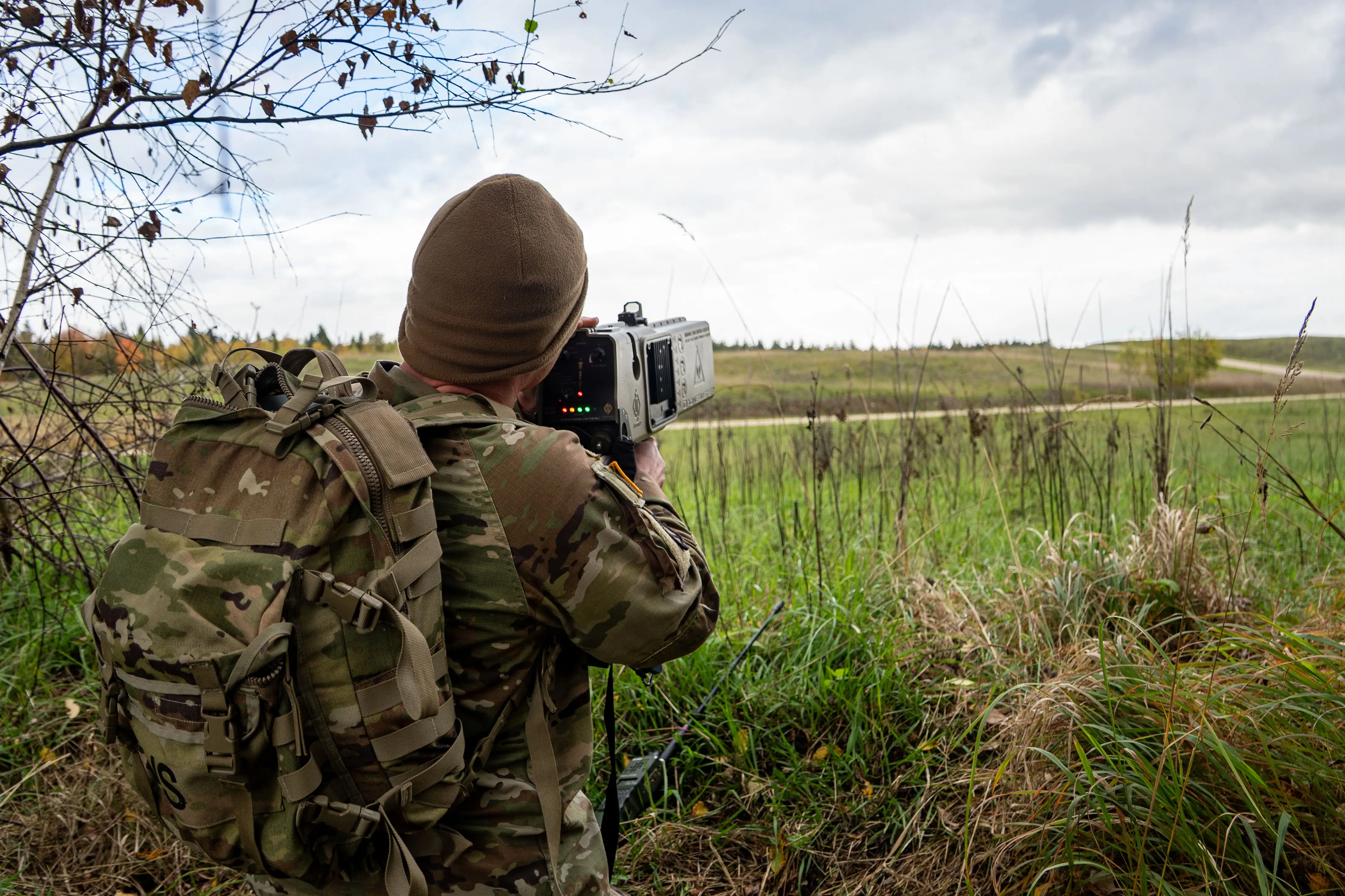A US Army soldier points in an area of grassy terrain with a Dronebuster.
