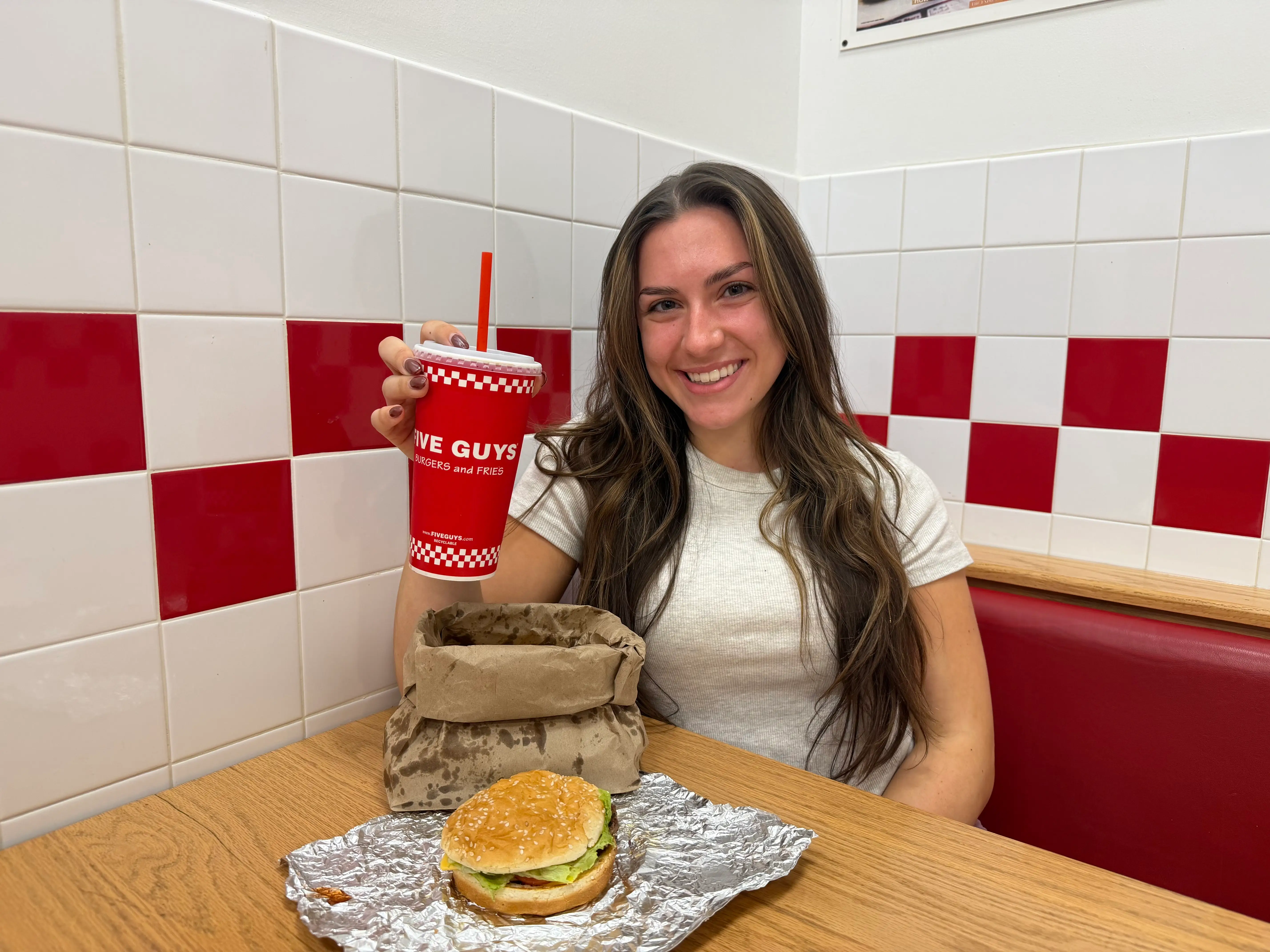 Amanda holds a soda and sits at a booth, with a cheeseburger and a bag of fries on the table.