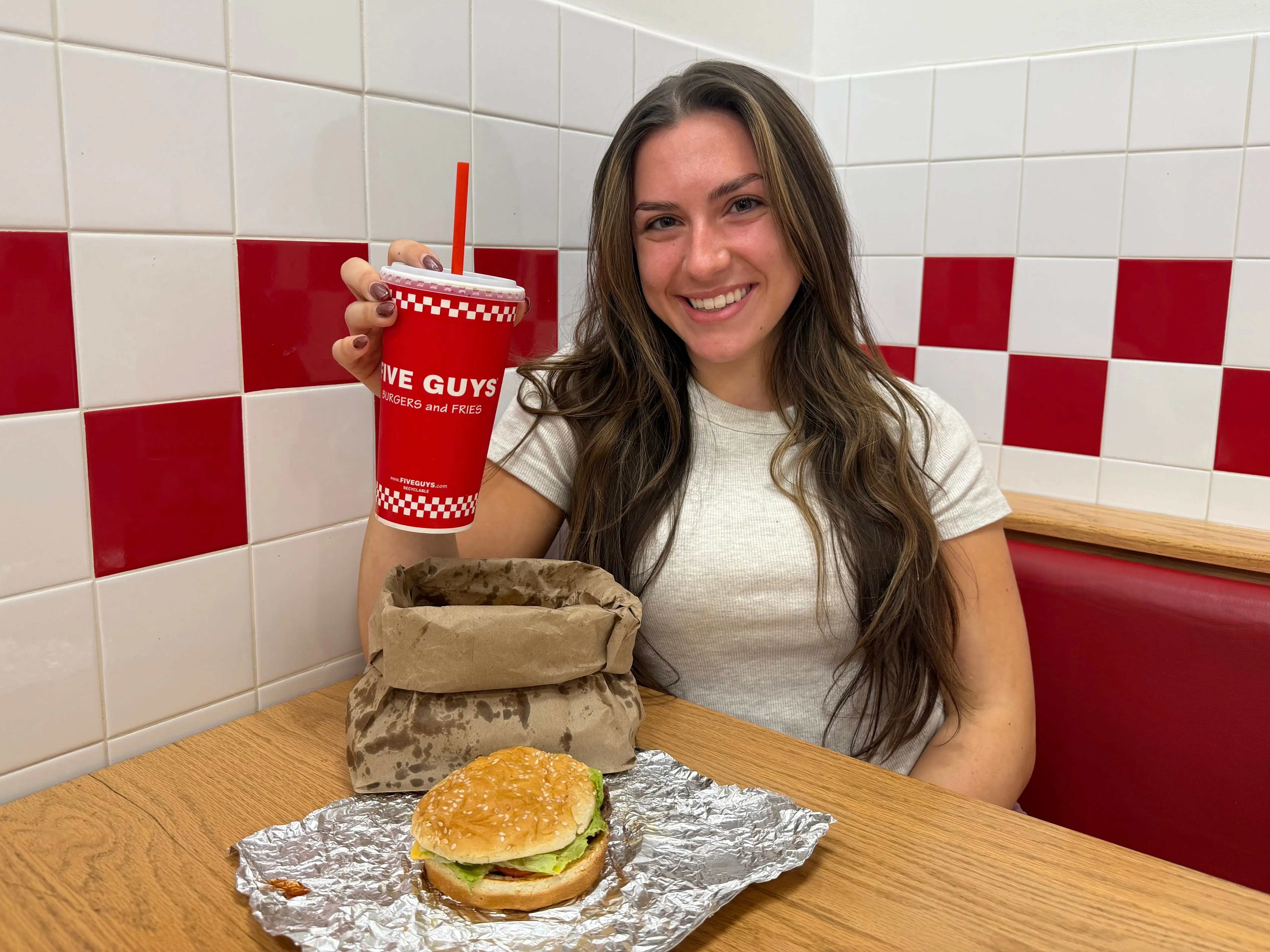 Amanda holds a soda and sits at a booth, with a cheeseburger and a bag of fries on the table.